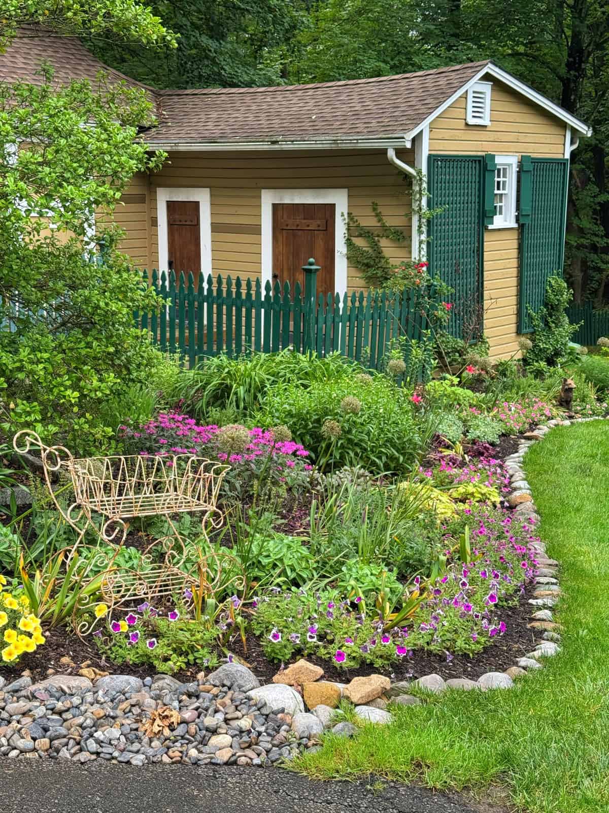A lush garden with blooming flowers and a decorative wire bench sits in front of a yellow shed with green shutters and a green picket fence, surrounded by trees and greenery.