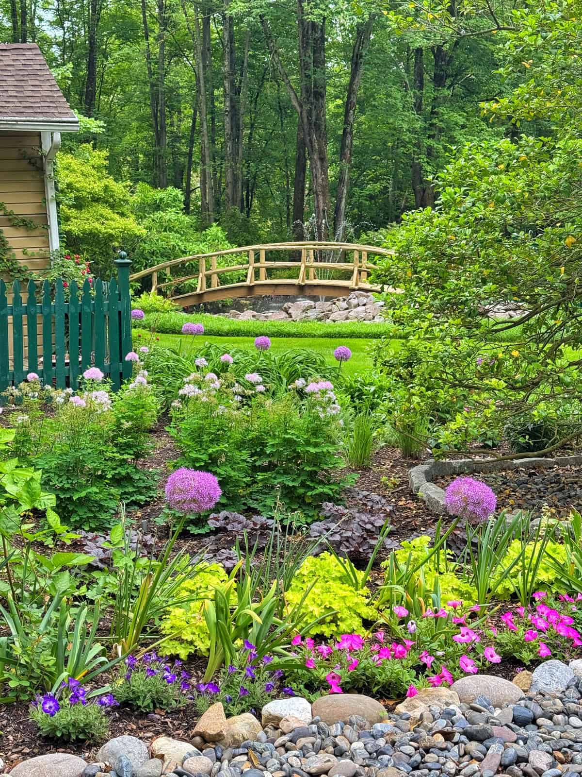 A lush garden with vibrant flowers, green shrubs, and purple blossoms in the foreground. A wooden fence and a small arching footbridge stand in the background, with tall trees and dense foliage beyond.