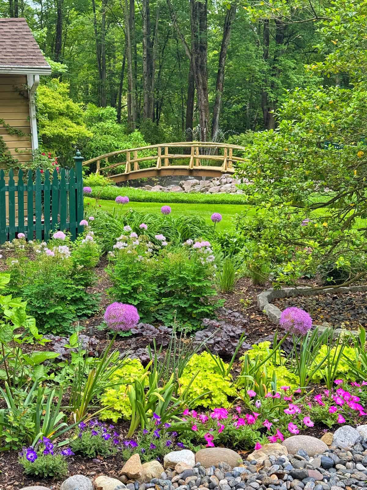 A lush garden with colorful flowers, green plants, and a small stone path in the foreground. A wooden footbridge crosses over a grassy area, and tall trees fill the background, creating a peaceful, scenic view.