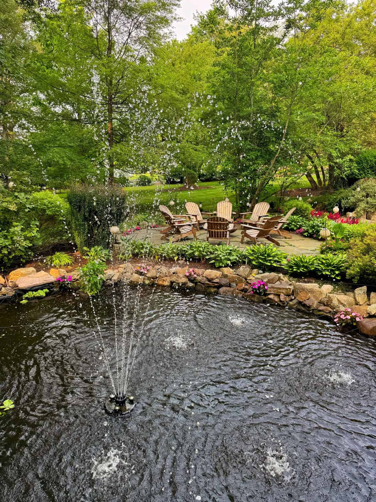 A fountain sprays water in the middle of a pond surrounded by rocks and flowers, with several wooden Adirondack chairs arranged on a stone patio in a lush, green garden.
