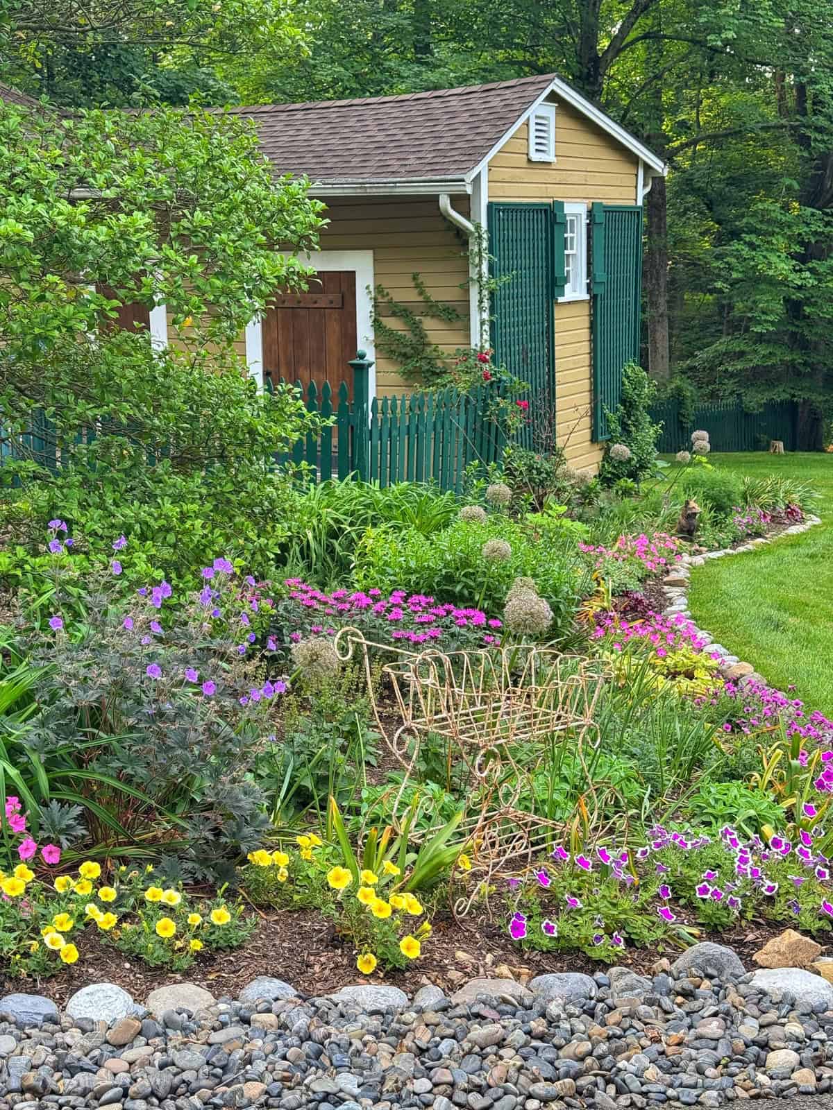 A lush garden with colorful flowers and a decorative metal bench sits in front of a yellow shed with green shutters and a fenced yard, bordered by a rock edge and surrounded by trees.