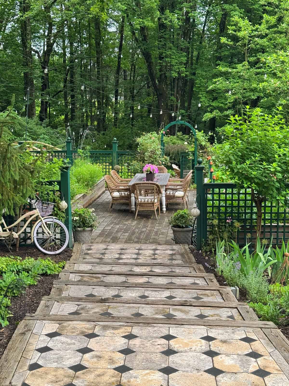 Stone steps lead to a garden patio with a table and wicker chairs. Pink flowers are arranged on the table, surrounded by lush greenery, trees, and a vintage bicycle with a basket of flowers. String lights hang overhead.