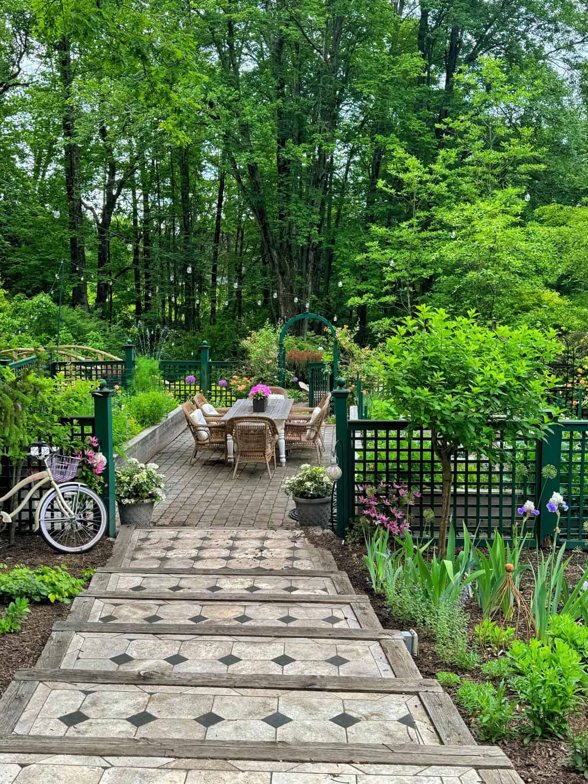 Stone steps lead down to a patio with a table and chairs, surrounded by lush greenery, blooming flowers, and a fenced garden area. A bicycle is parked to the left, and tall trees provide a serene backdrop.
