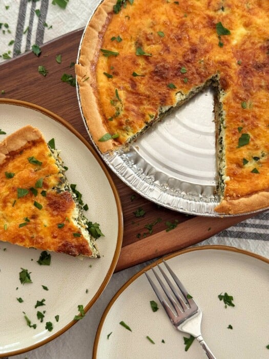 A baked ricotta and spinach quiche with a golden crust sits on a board, with one slice removed and placed on a plate nearby. Chopped herbs are sprinkled on the quiche and plate, and a fork rests on a second empty plate.