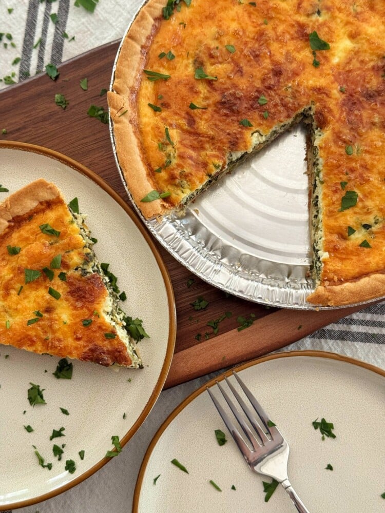 A baked ricotta and spinach quiche with a golden crust sits on a board, with one slice removed and placed on a plate nearby. Chopped herbs are sprinkled on the quiche and plate, and a fork rests on a second empty plate.