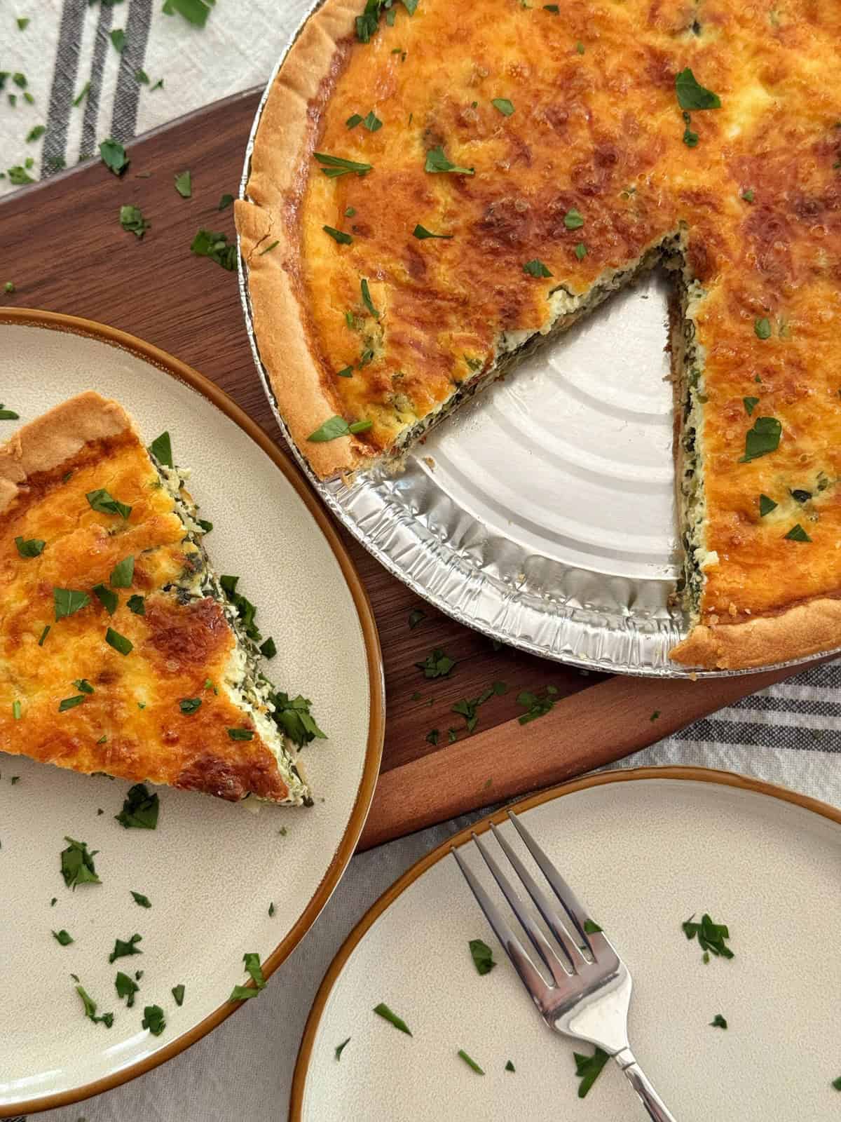 A baked ricotta and spinach quiche with a golden crust sits on a board, with one slice removed and placed on a plate nearby. Chopped herbs are sprinkled on the quiche and plate, and a fork rests on a second empty plate.