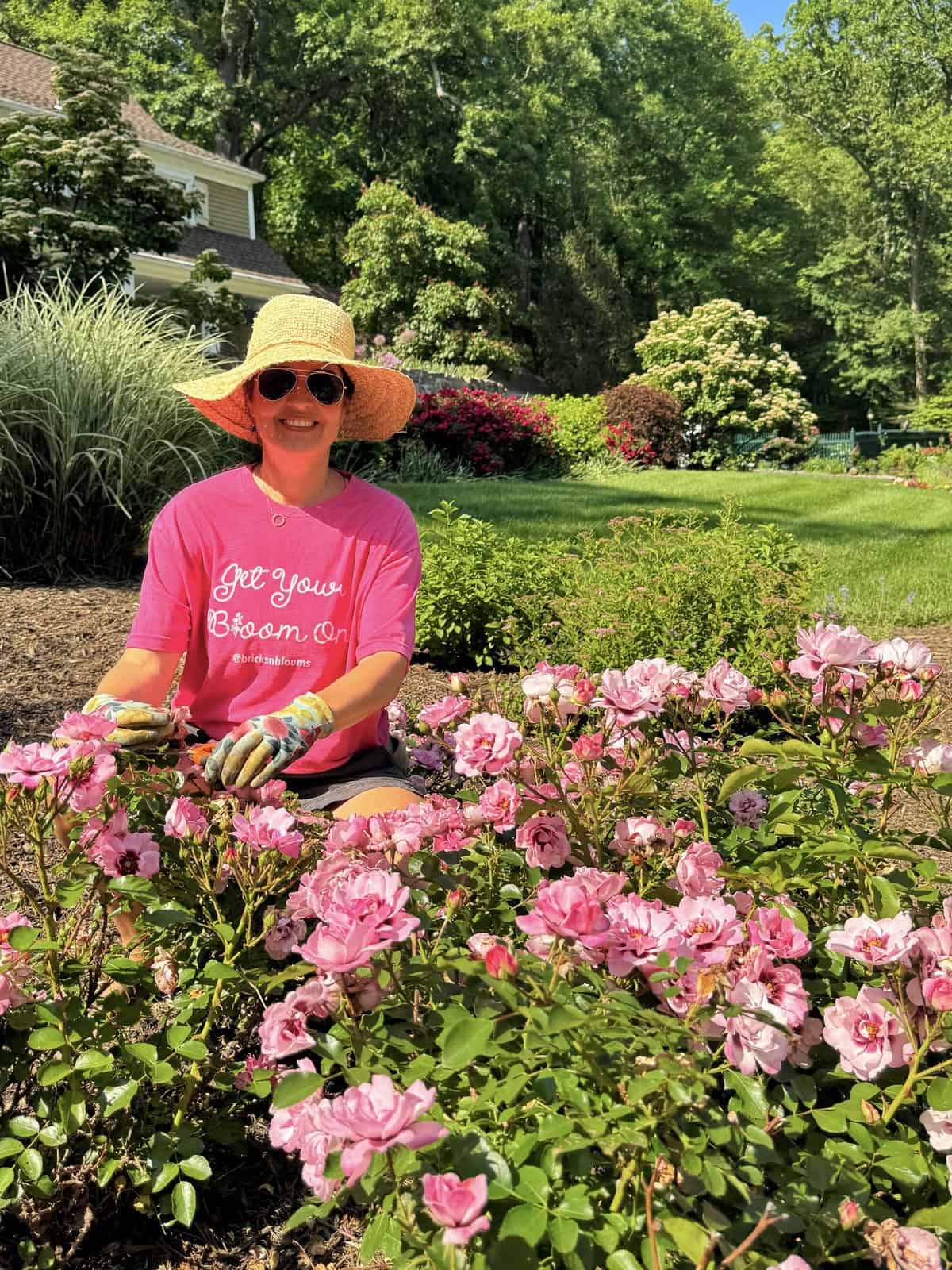 A woman wearing a straw hat, sunglasses, gloves, and a pink shirt sits in a garden, smiling while tending to blooming pink flowers on a sunny day. Lush greenery and a house are visible in the background.