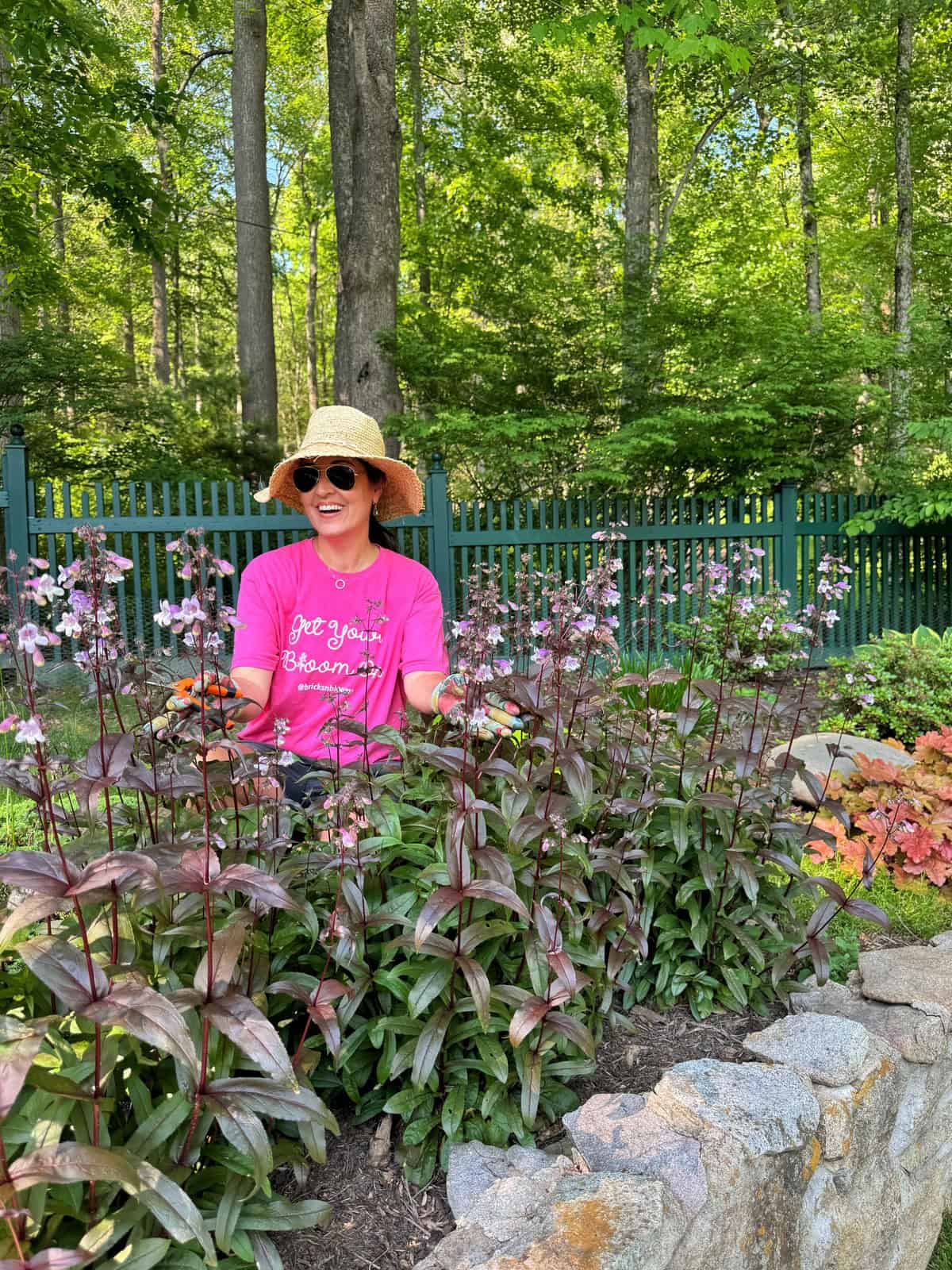 A woman wearing a straw hat, sunglasses, and a pink shirt sits in a garden behind tall flowering plants, smiling. A stone border and green fence are in the background amid lush trees.