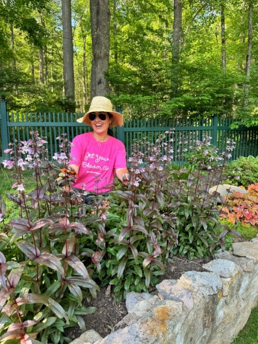 A smiling person wearing a straw hat, sunglasses, and a bright pink shirt sits among blooming flowers in a garden, with a green fence and trees in the background.