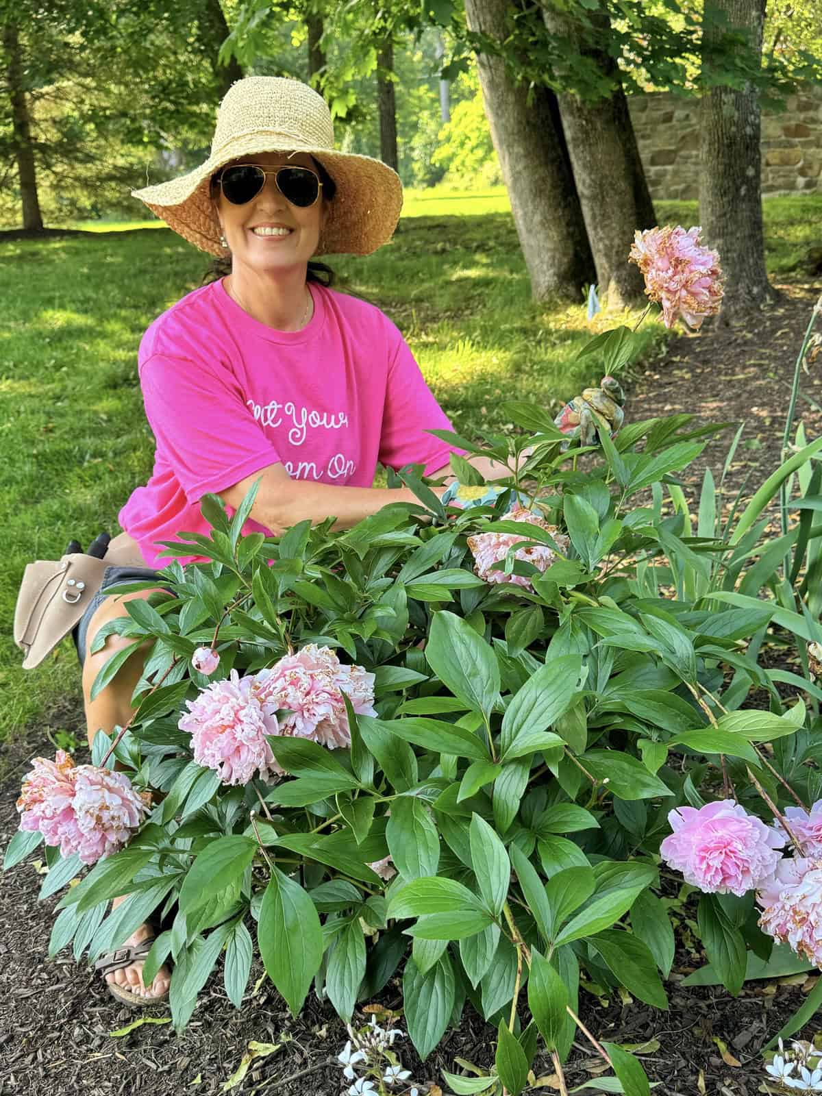 A smiling woman in a pink shirt, sun hat, and sunglasses kneels in a garden beside blooming pink peonies. Tall trees and a stone wall are visible in the sunlit background.