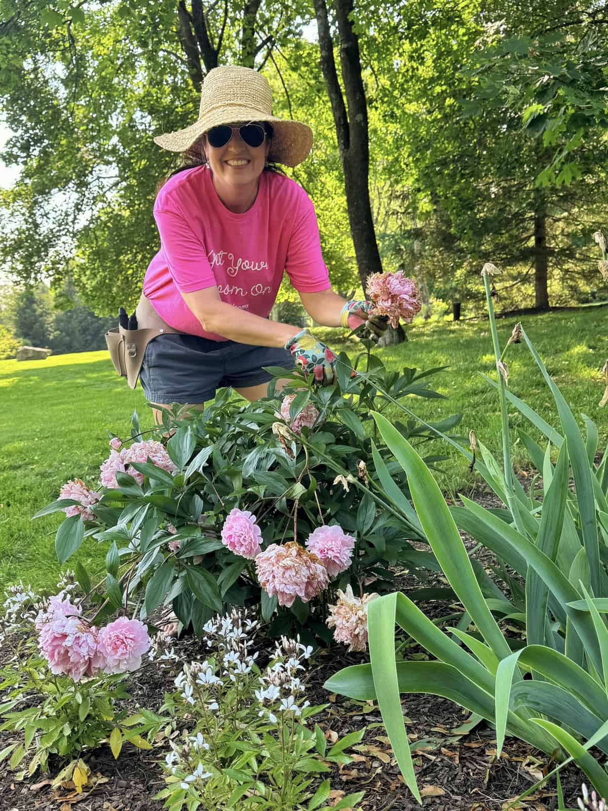 A smiling person in a pink shirt, straw hat, and sunglasses tends to blooming pink flowers in a sunny garden with green trees and grass in the background.