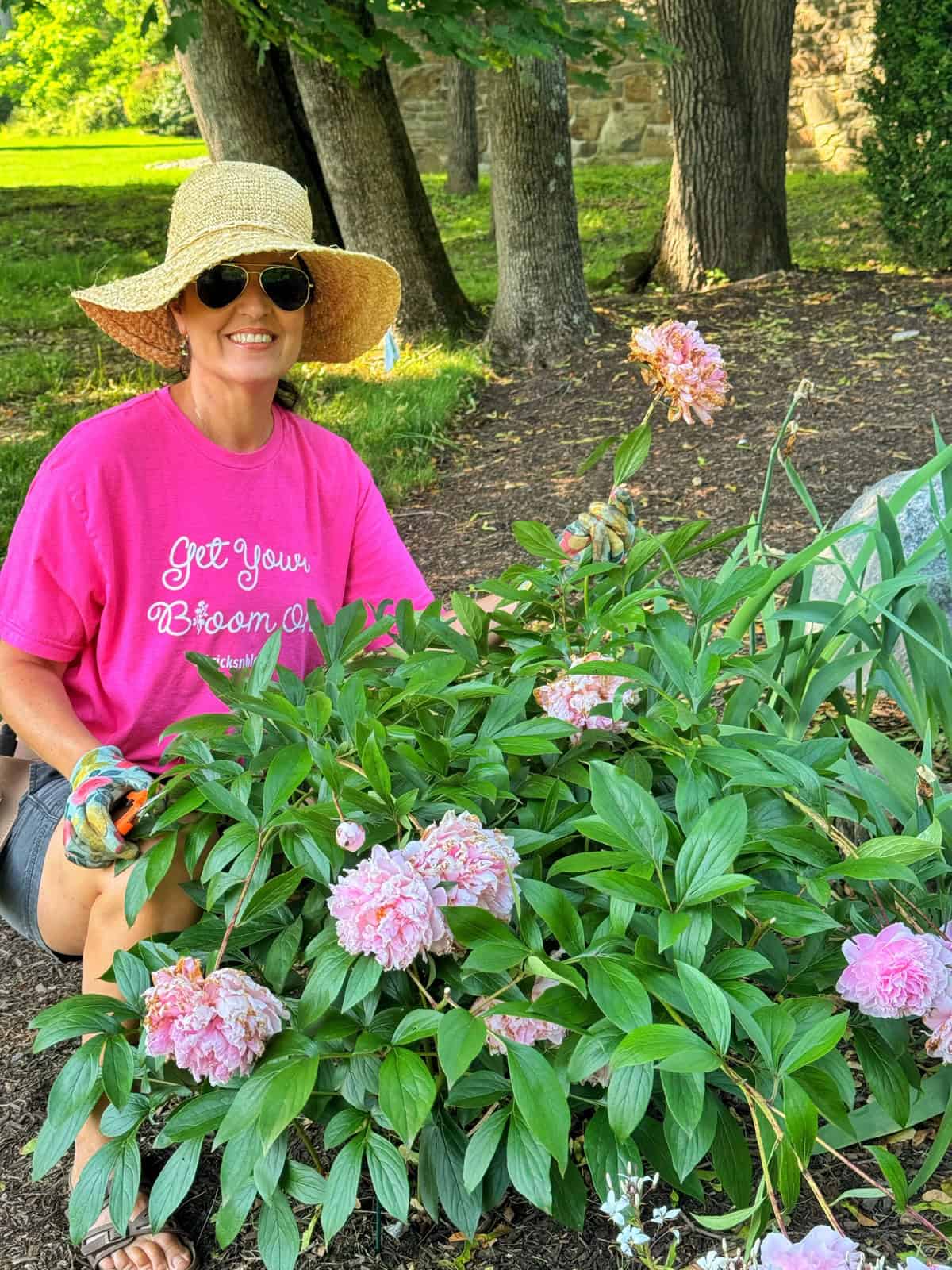 A smiling woman wearing a straw hat, sunglasses, pink shirt, and gloves kneels in a garden, pruning blooming pink peonies. Trees and greenery are visible in the background.