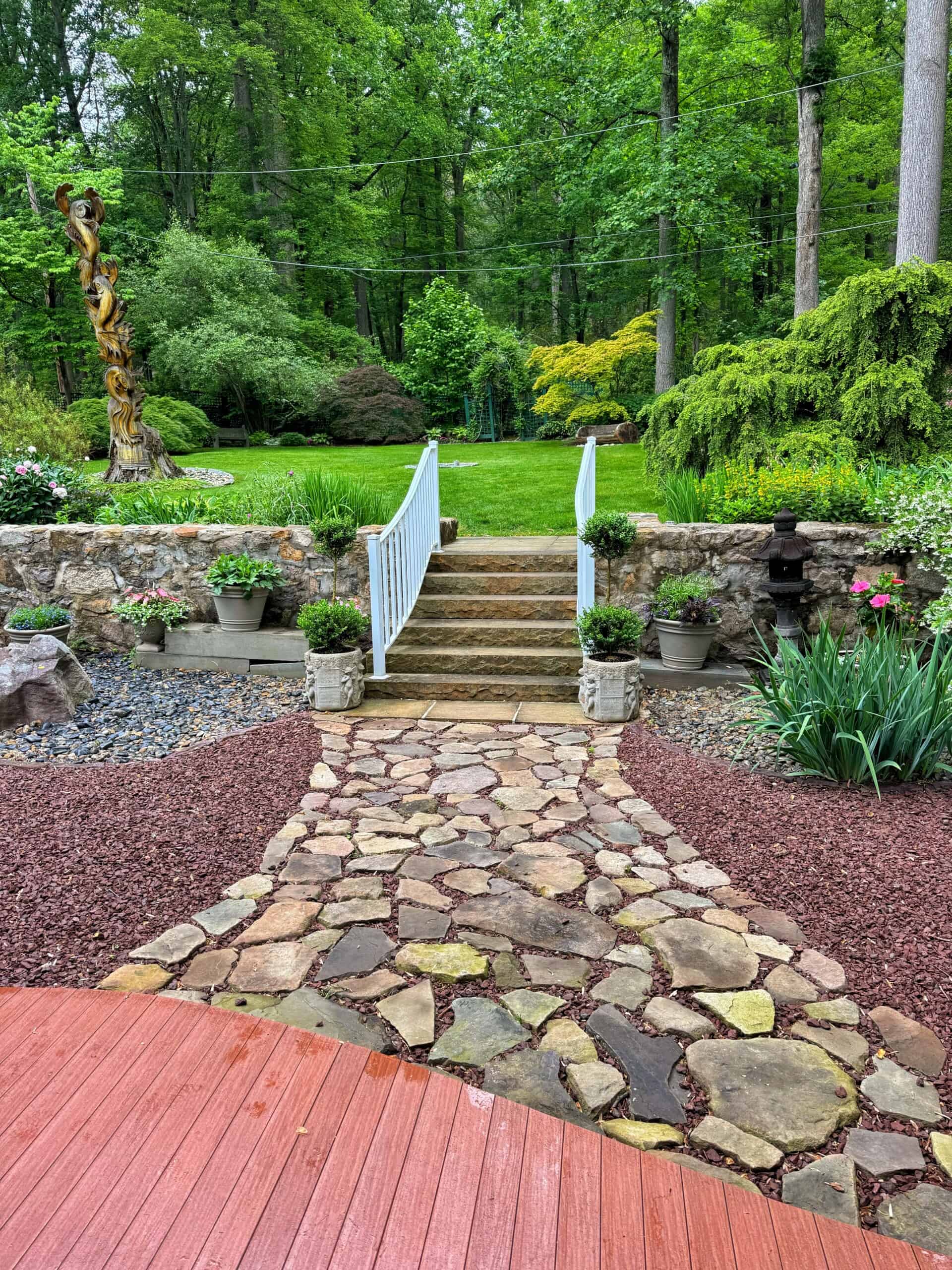 A stone pathway leads from a red deck to steps with white railings, flanked by potted plants, ending at a large green lawn bordered by trees and landscaped garden beds.