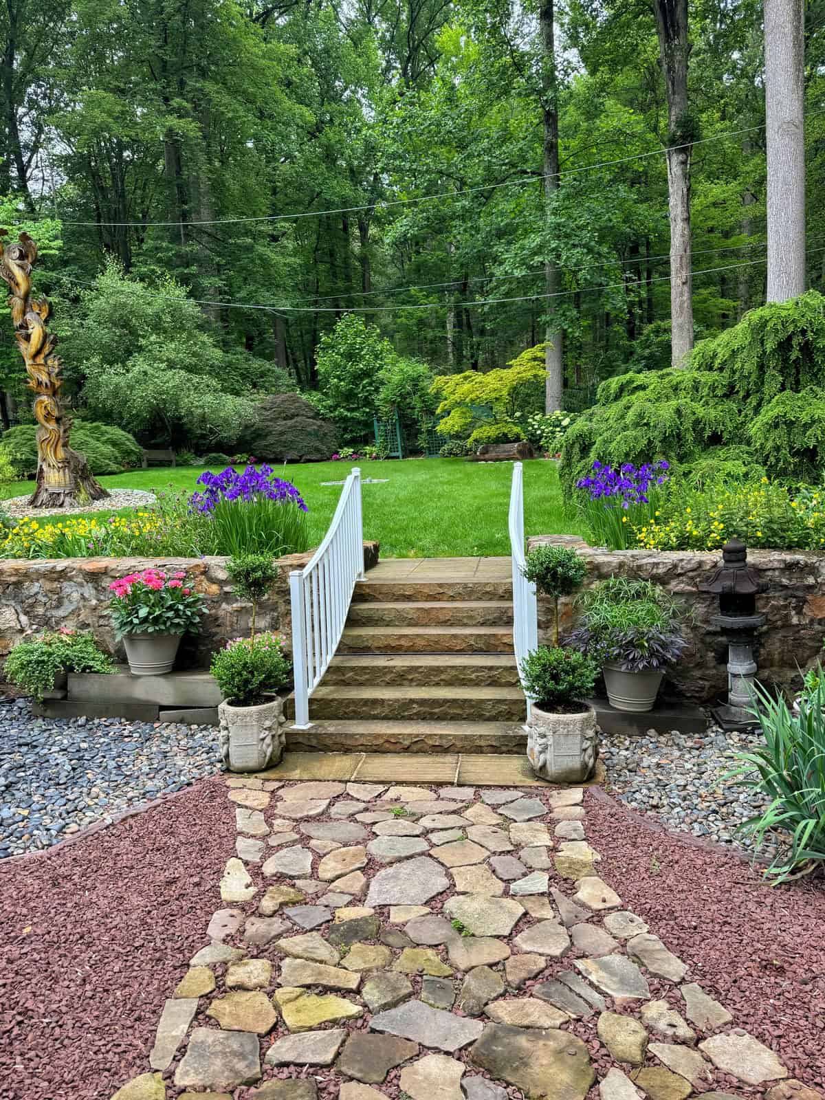 A stone path leads to steps with a white railing, surrounded by potted flowers and lush greenery, opening into a well-kept lawn bordered by trees and flowering plants in a peaceful garden setting.
