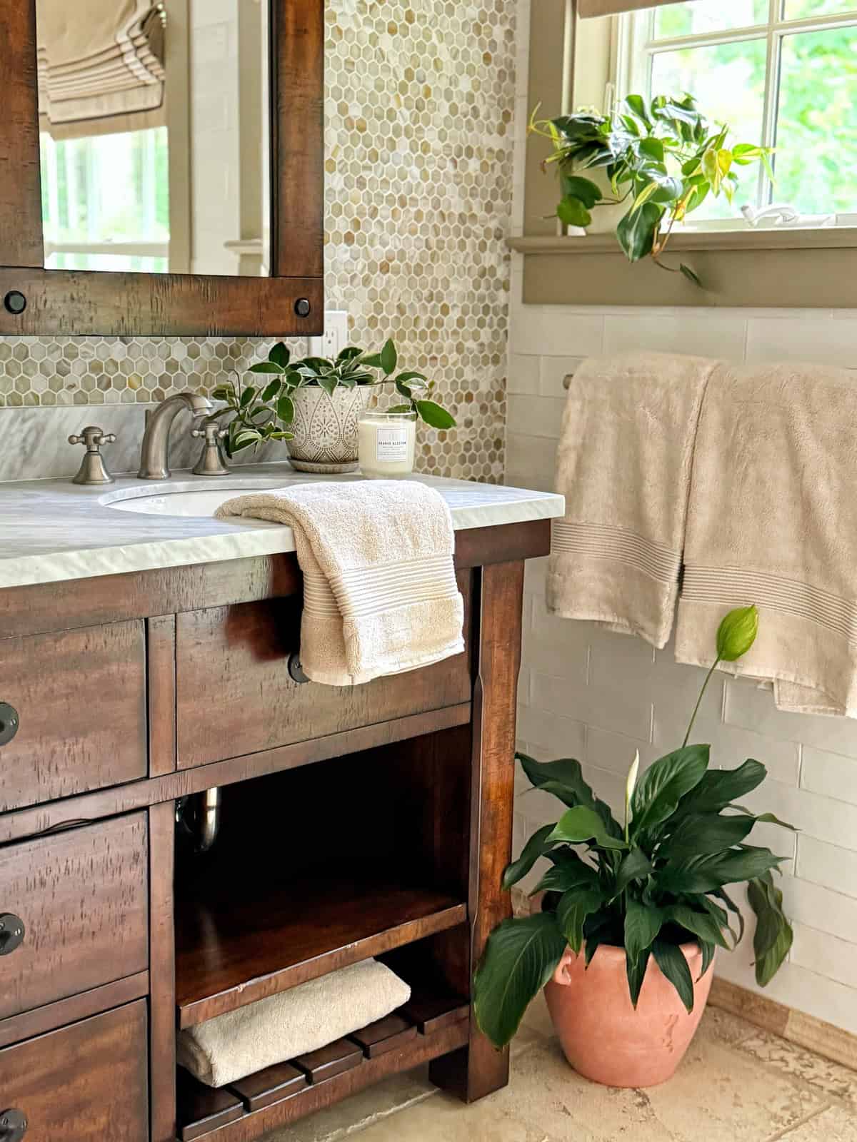 A bathroom with a wooden vanity, white countertop, and potted plants. Beige towels hang on a rack and are folded on the shelf. Sunlight streams through a window above the sink, highlighting the clean, natural decor.