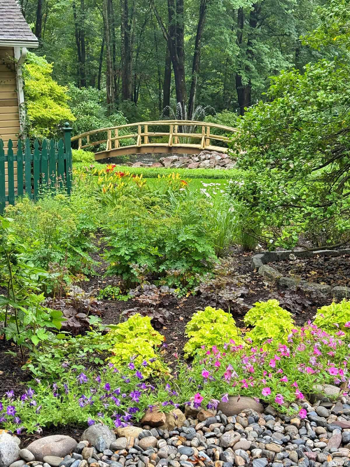 A lush garden with colorful flowers, green shrubs, and a stone path, leading to a small wooden footbridge over a stream; trees and a green fence are in the background.