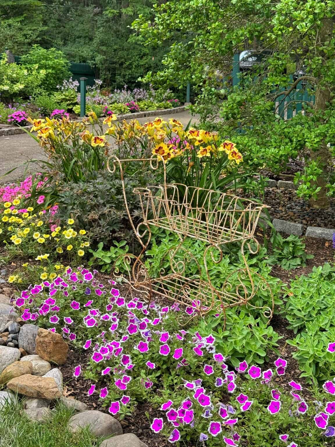 A decorative metal bench sits in a lush garden bed filled with bright yellow, orange, and pink flowers, surrounded by green foliage and bordered by rocks with a pathway and more greenery in the background.