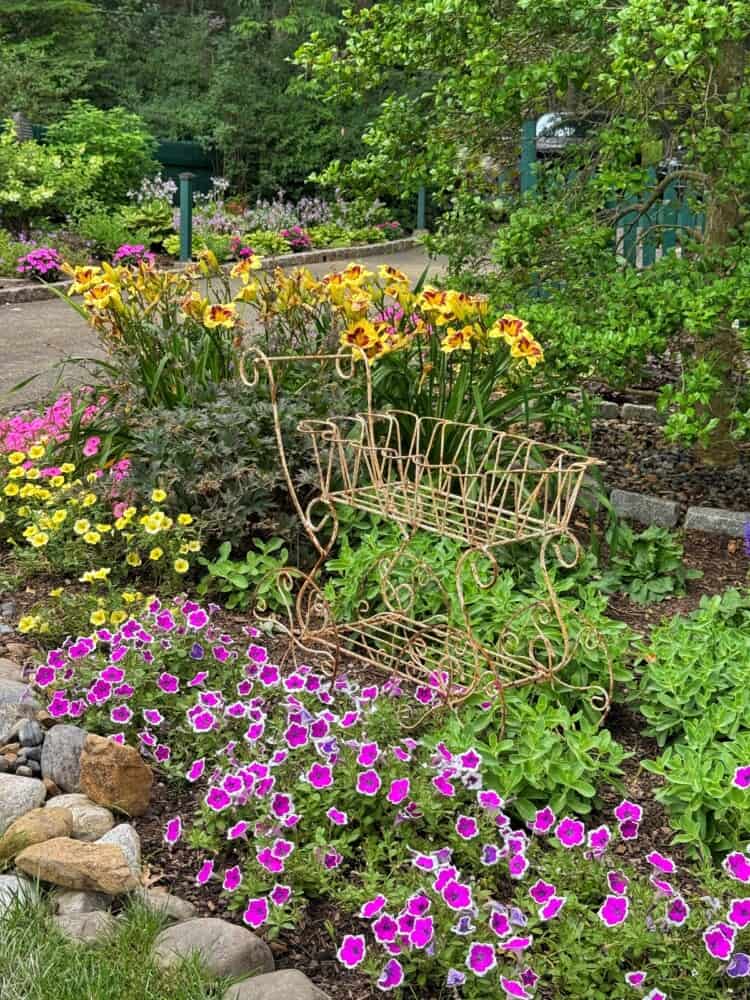 A decorative metal bench sits in a lush garden bed filled with bright yellow, orange, and pink flowers, surrounded by green foliage and bordered by rocks with a pathway and more greenery in the background.