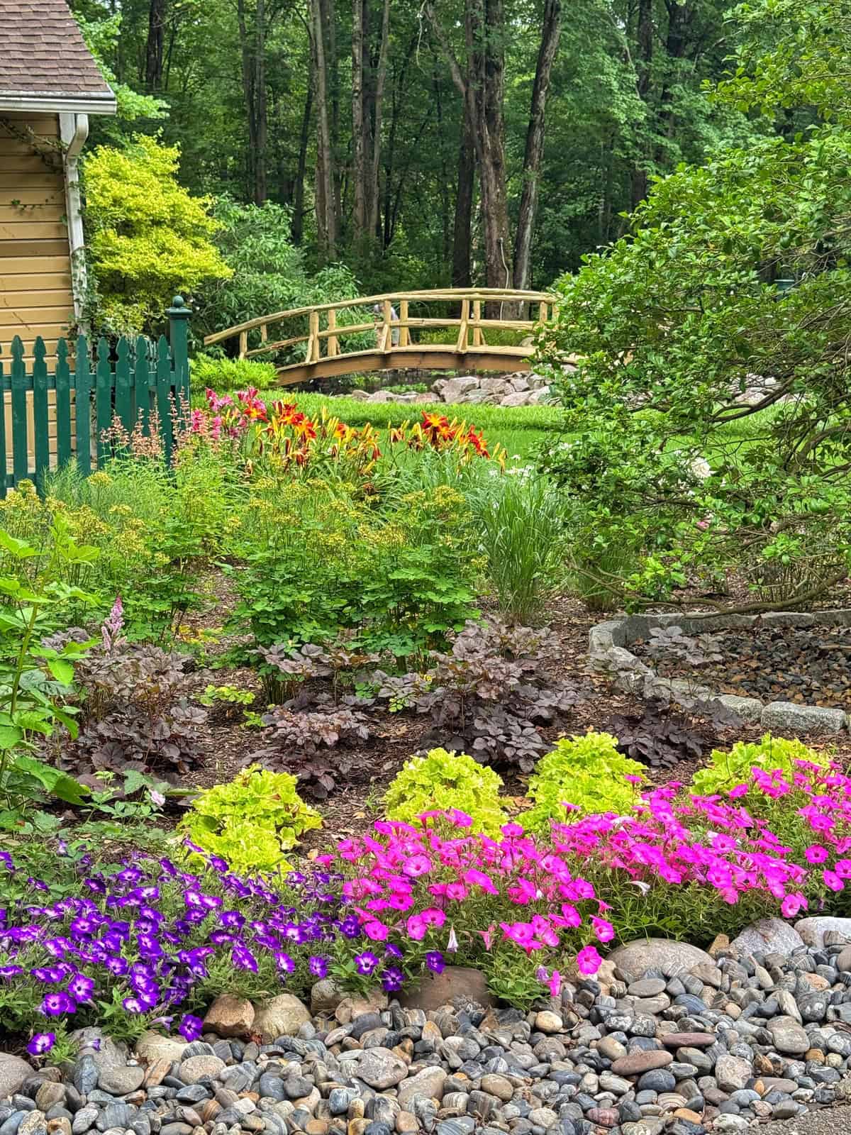 A garden with colorful flowers, green shrubs, and a rocky path. In the background, there is a yellow picket fence, a small wooden bridge, and tall trees.