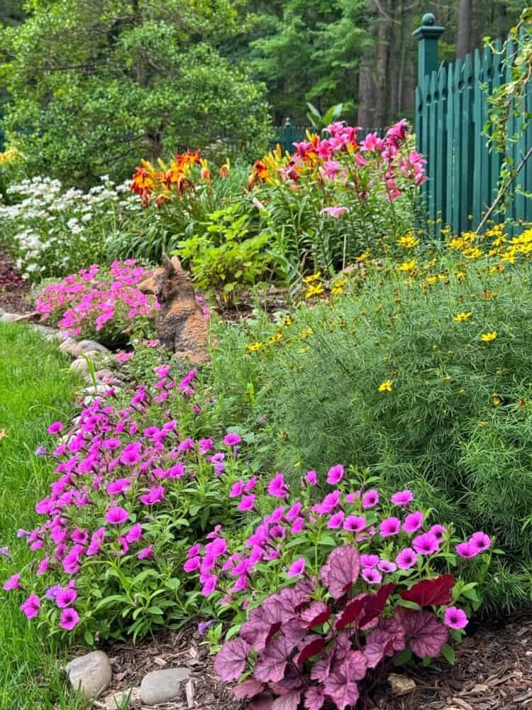 A colorful cottage garden with blooming pink, orange, yellow, and white flowers, dense green foliage, and a green fence in the background. A small brown statue of an animal sits among the flowers.