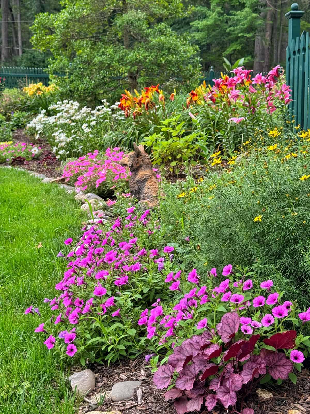 A lush flower garden with vibrant pink, orange, yellow, and white flowers; a stone border separates the flowers from the green grass. A small, fluffy brown cat sits among the blooms. Green fencing and trees are in the background.