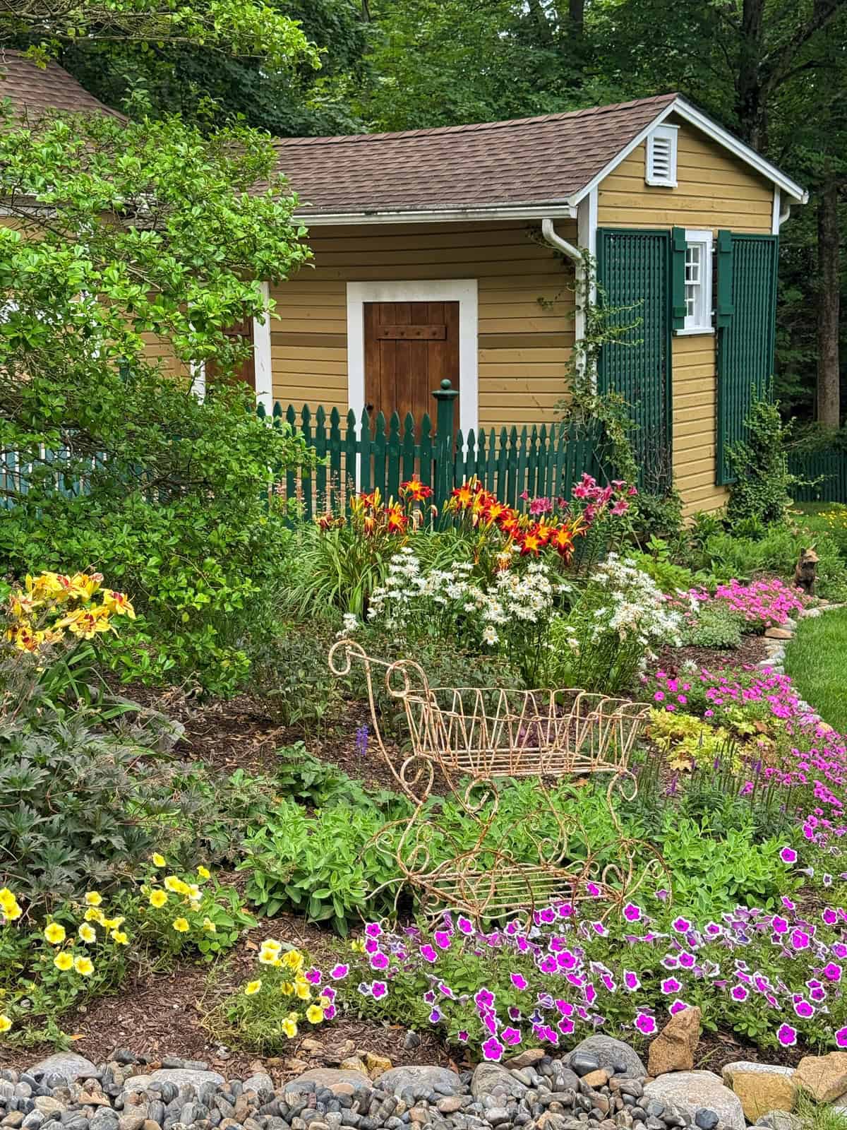 A lush garden with vibrant flowers, including lilies and petunias, surrounds a decorative metal bench. In the background, there is a yellow garden shed with green trim and a picket fence. Rocks border the flower bed.
