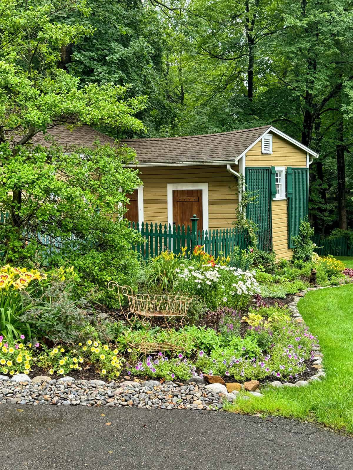 Yellow garden shed with green shutters and a green picket fence, surrounded by lush trees and vibrant flower beds featuring a variety of colorful blooms, next to a neatly maintained lawn and stone-bordered garden.