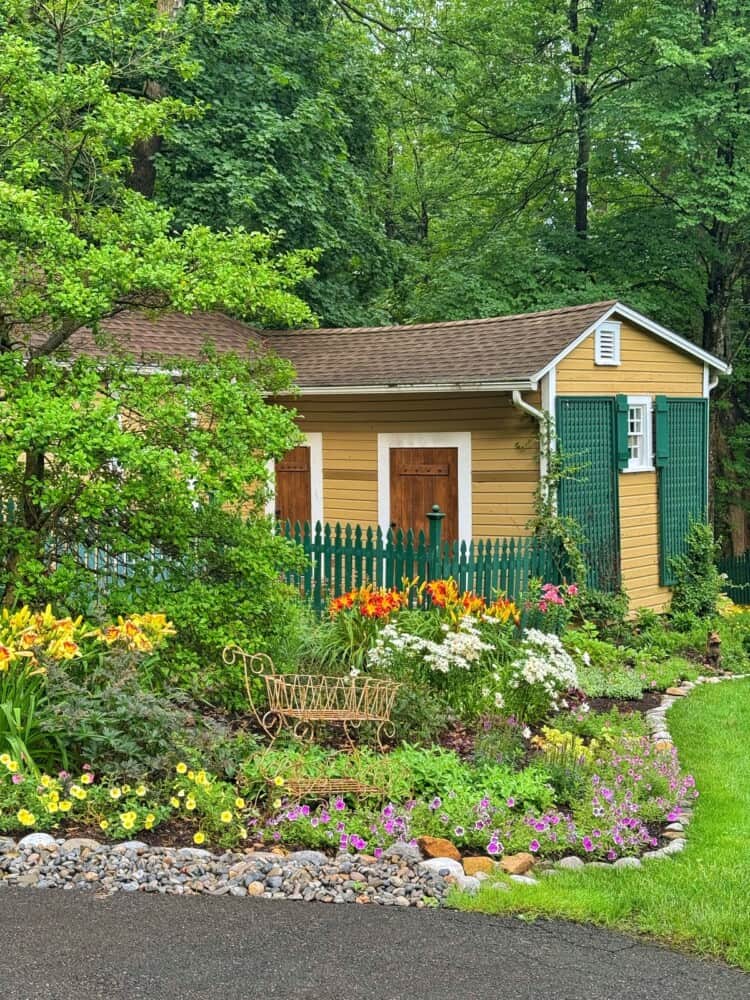 A small yellow garden shed with brown doors sits behind a green picket fence, surrounded by lush plants, colorful flowers, and a decorative metal bench in a well-maintained garden. Trees provide a green backdrop.