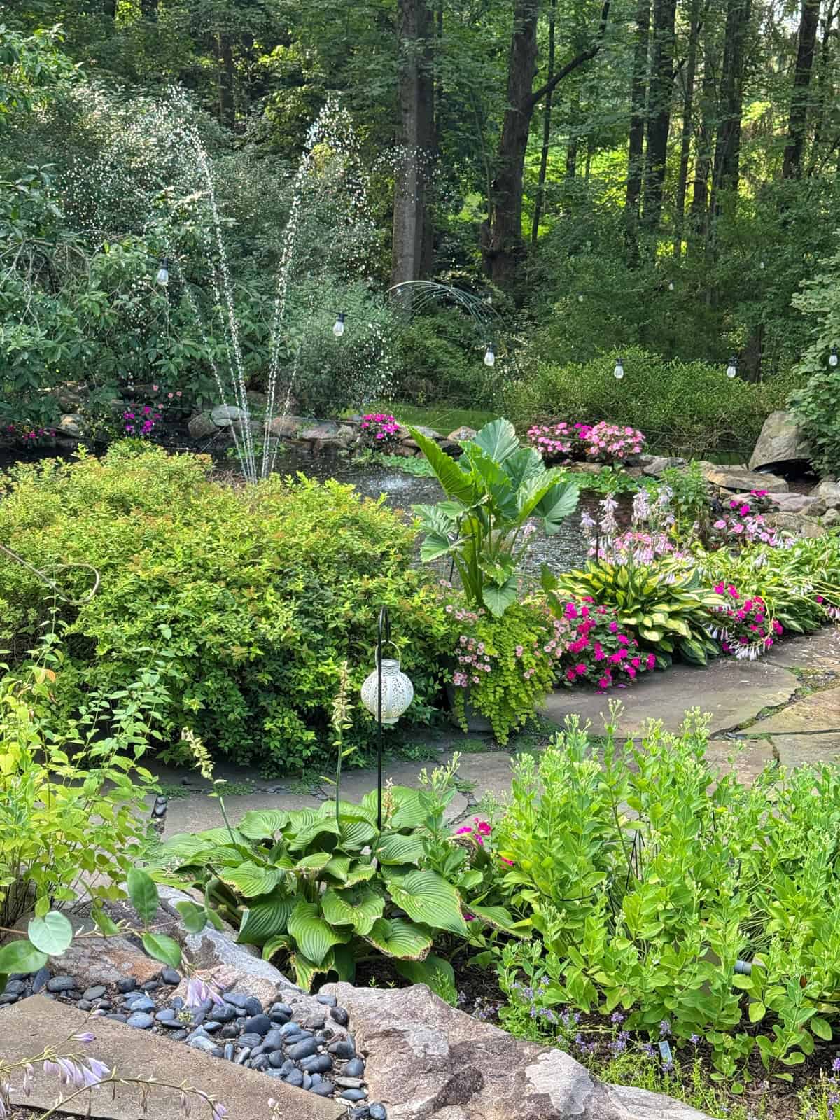 A lush garden with vibrant green plants, blooming pink and purple flowers, a stone path, and a water fountain spraying arcs of water. Strings of lights and tall trees create a peaceful, shaded woodland setting.