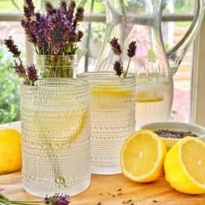 Two textured glasses of lavender lemonade garnished with lavender sit on a wooden board with a halved lemon, loose lavender sprigs, and a glass pitcher of lemonade, with lavender flowers in a jar in the background.