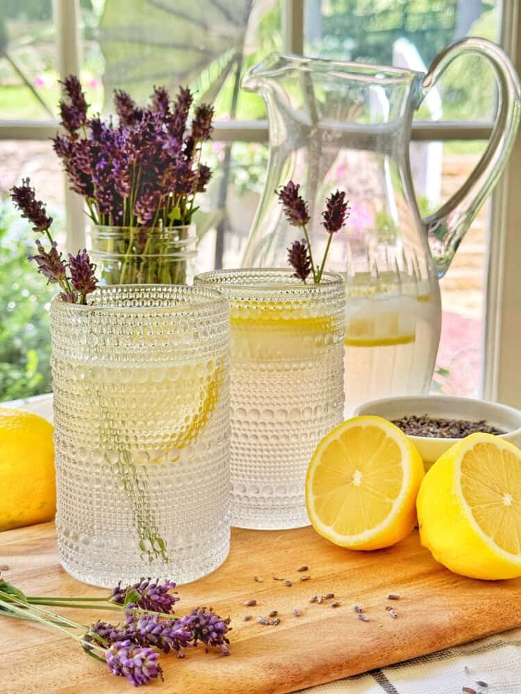 Two textured glasses of lavender lemonade garnished with lavender sit on a wooden board with a halved lemon, loose lavender sprigs, and a glass pitcher of lemonade, with lavender flowers in a jar in the background.