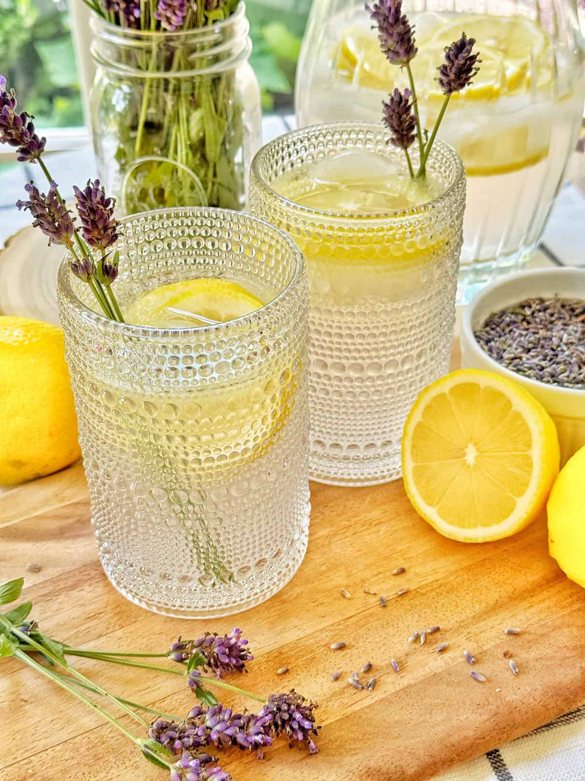 Two textured glasses of lavender lemonade with ice, lemon slices, and sprigs of lavender on a wooden board. Surrounding them are fresh lemons, a bowl of dried lavender, and a jar of lavender stems in the background.