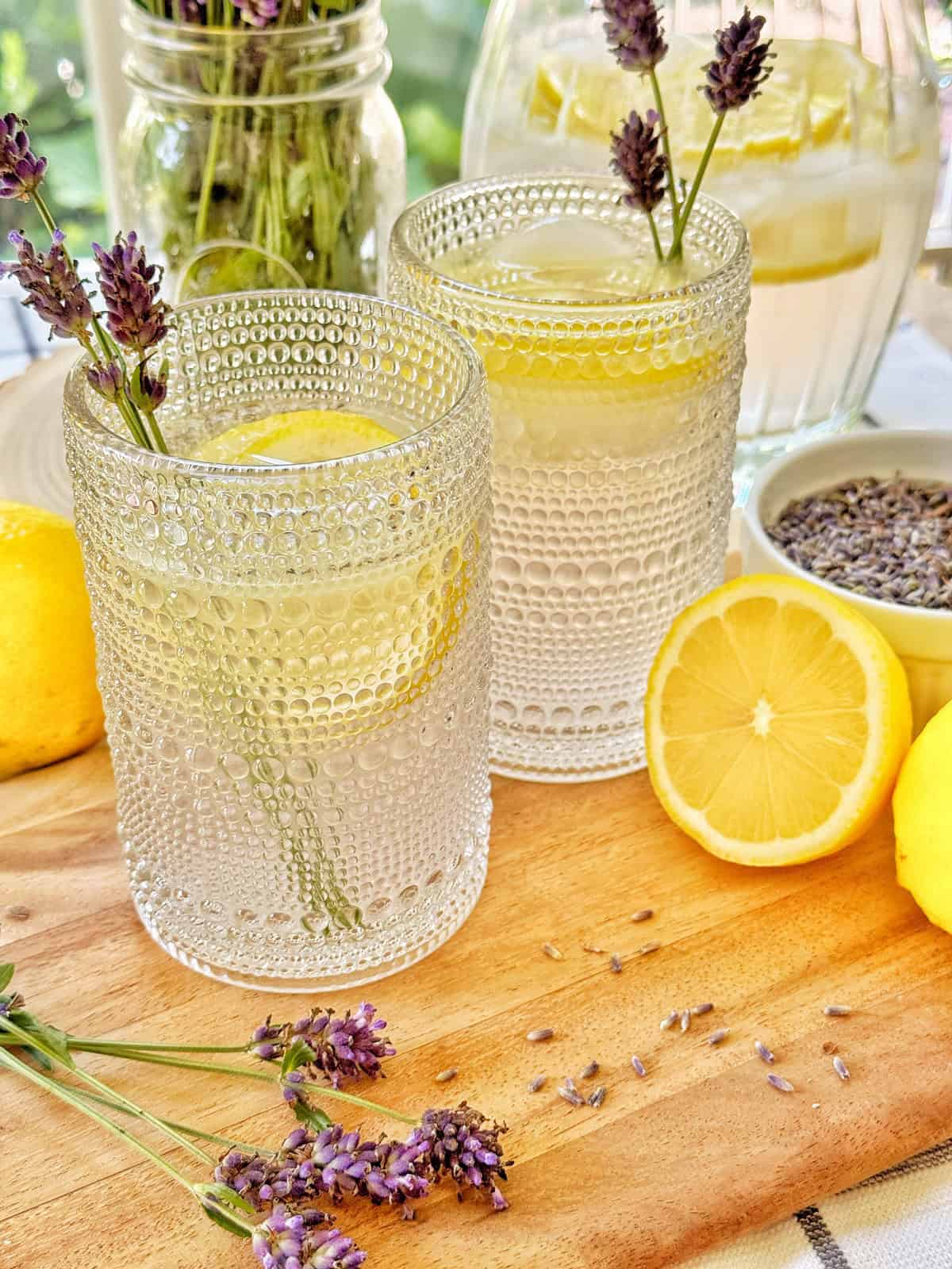 Two textured glasses filled with lemon water are garnished with lemon slices and lavender sprigs. Fresh lemons, a bowl of dried lavender, and a jar and pitcher of lemon water are arranged on a wooden surface.