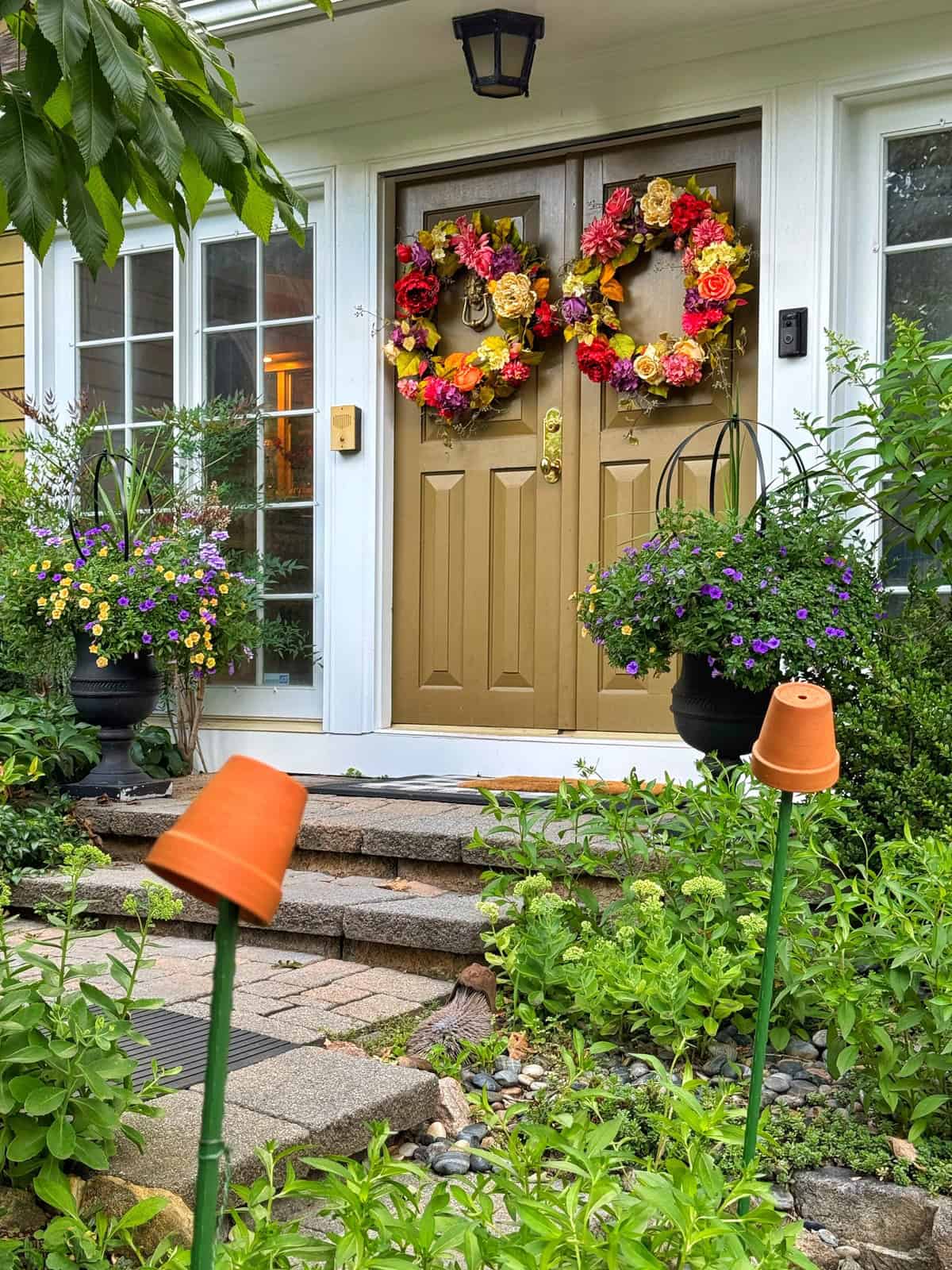A front door with two yellow wreaths made of colorful flowers, flanked by large potted plants with purple and yellow blooms. Clay pots are placed upside down on garden stakes along the stone path.