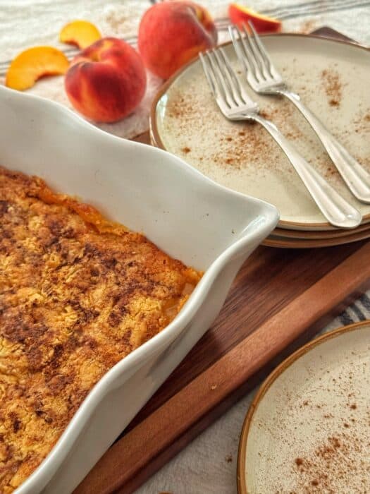 A peach dump cake recipe in a white baking dish sits on a wooden board beside stacked plates, three forks, and fresh peaches, with a sprinkle of cinnamon on the plates.