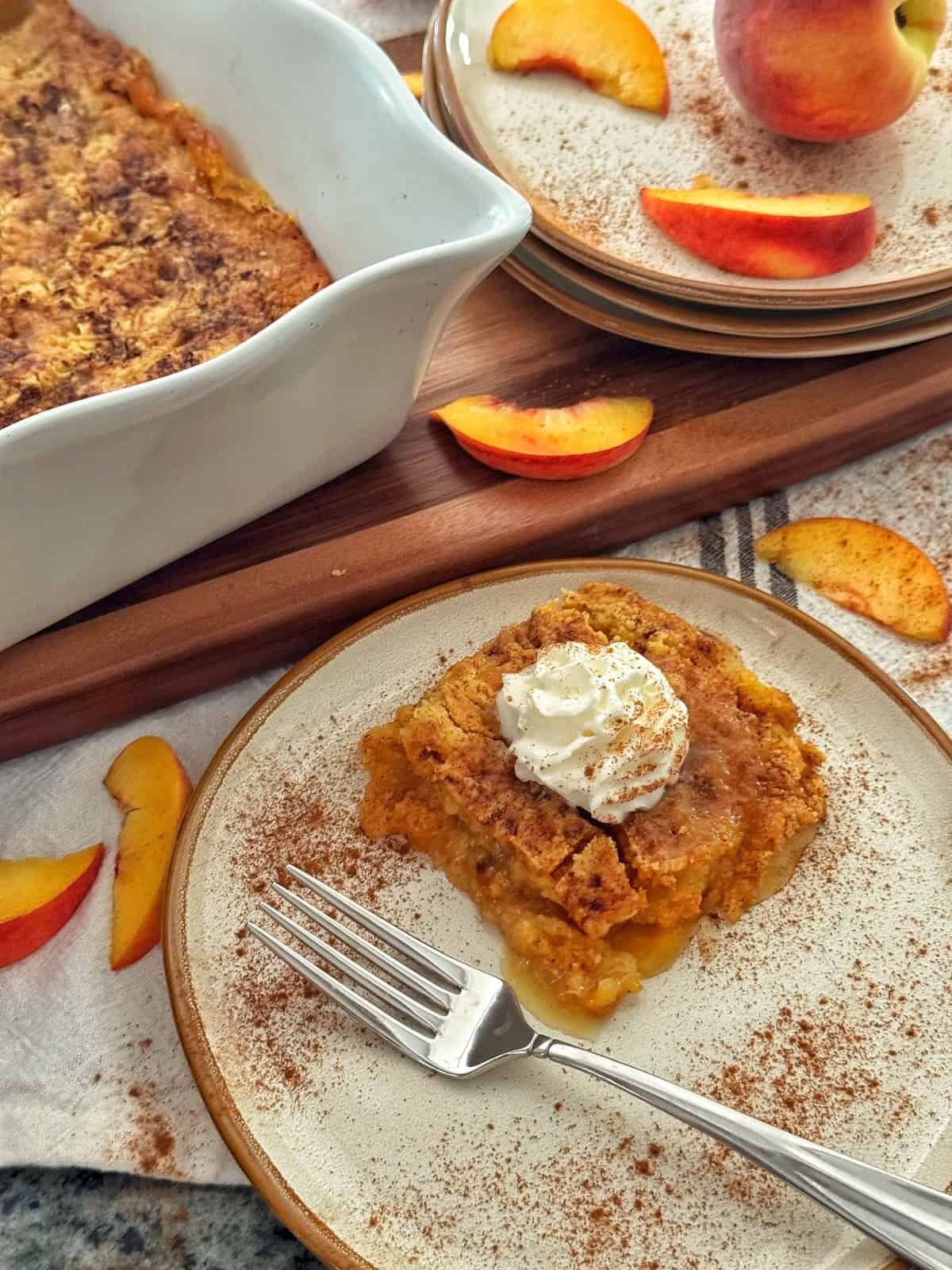 A slice of peach dump cake topped with whipped cream and cinnamon sits on a plate with a fork. Nearby are peach slices, a casserole dish of cobbler, and stacked plates, all set on a wooden surface.