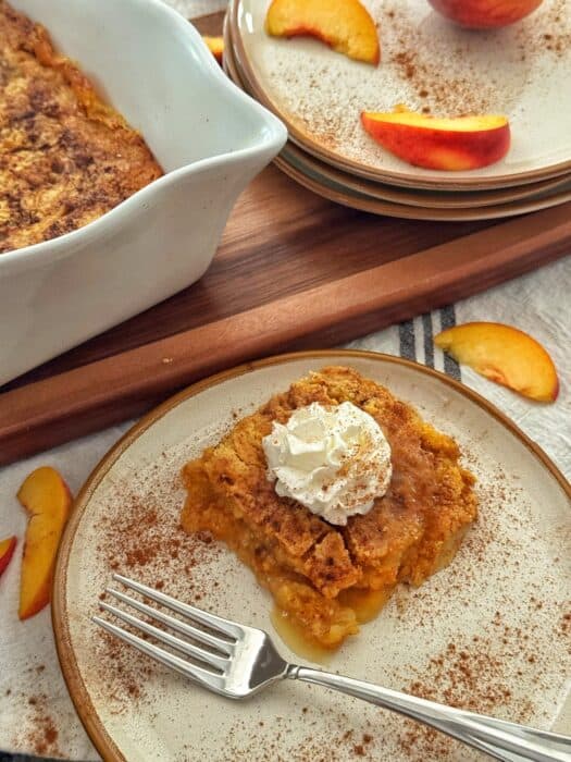 A slice of recipe for peach dump cake topped with whipped cream sits on a plate with a fork. There are cinnamon sprinkles and peach slices nearby, and more cobbler and stacked plates are in the background.