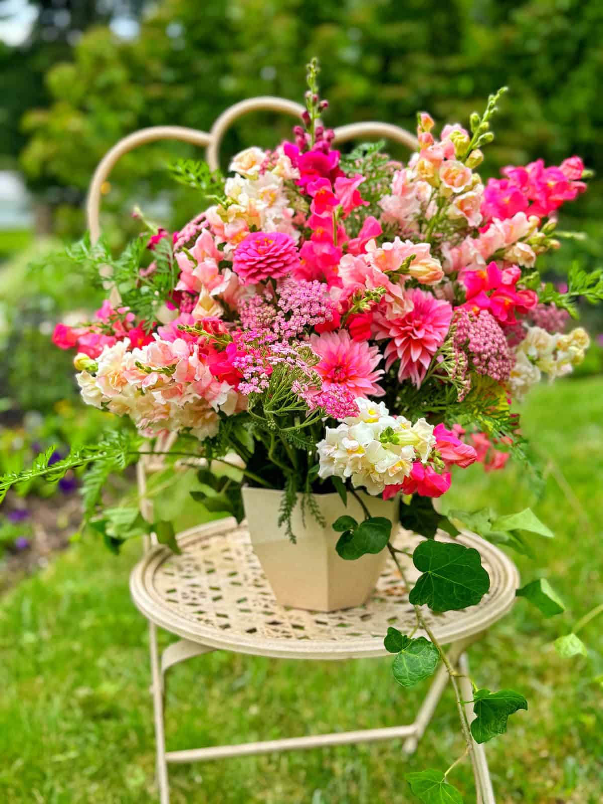 A vibrant bouquet of pink, white, and peach flowers arranged in a beige square pot sits on a woven chair outdoors, with green foliage in the background.