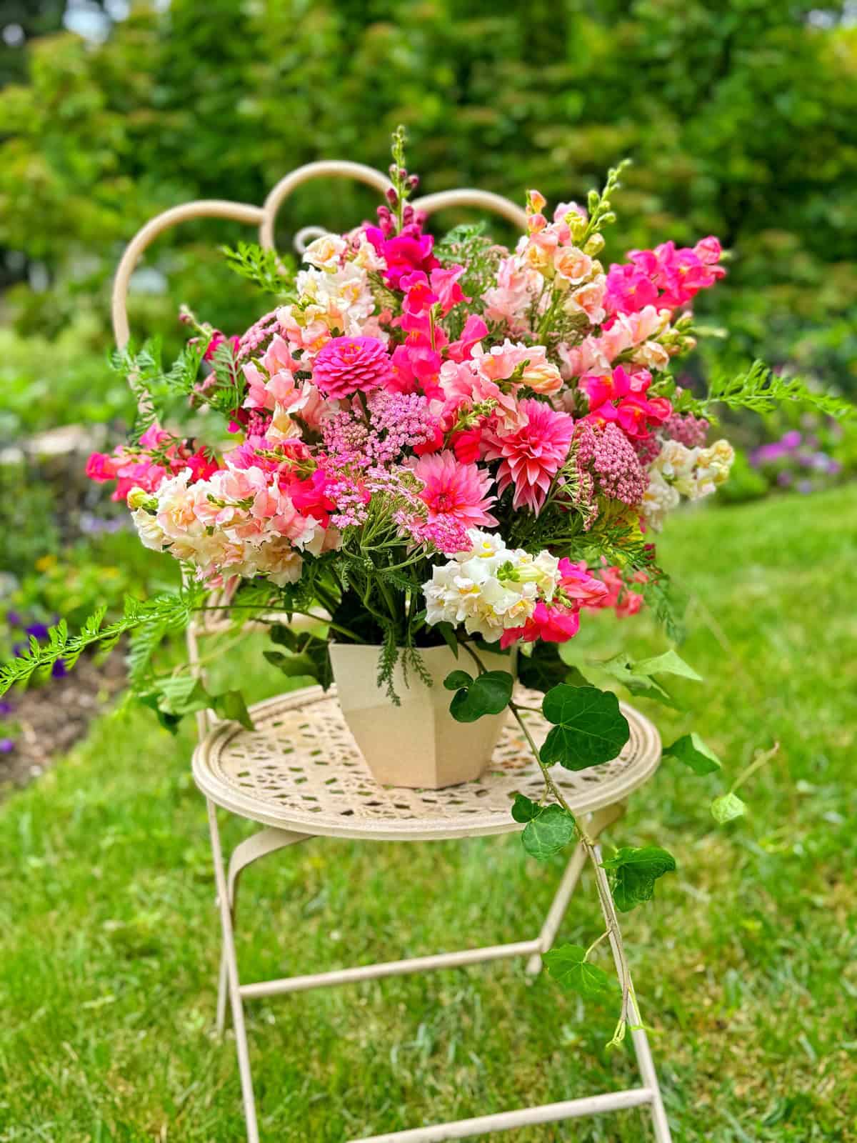 A pale metal chair sits on grass, holding a cream vase filled with a lush bouquet of pink, peach, and white flowers and green foliage, with a garden blurred in the background.