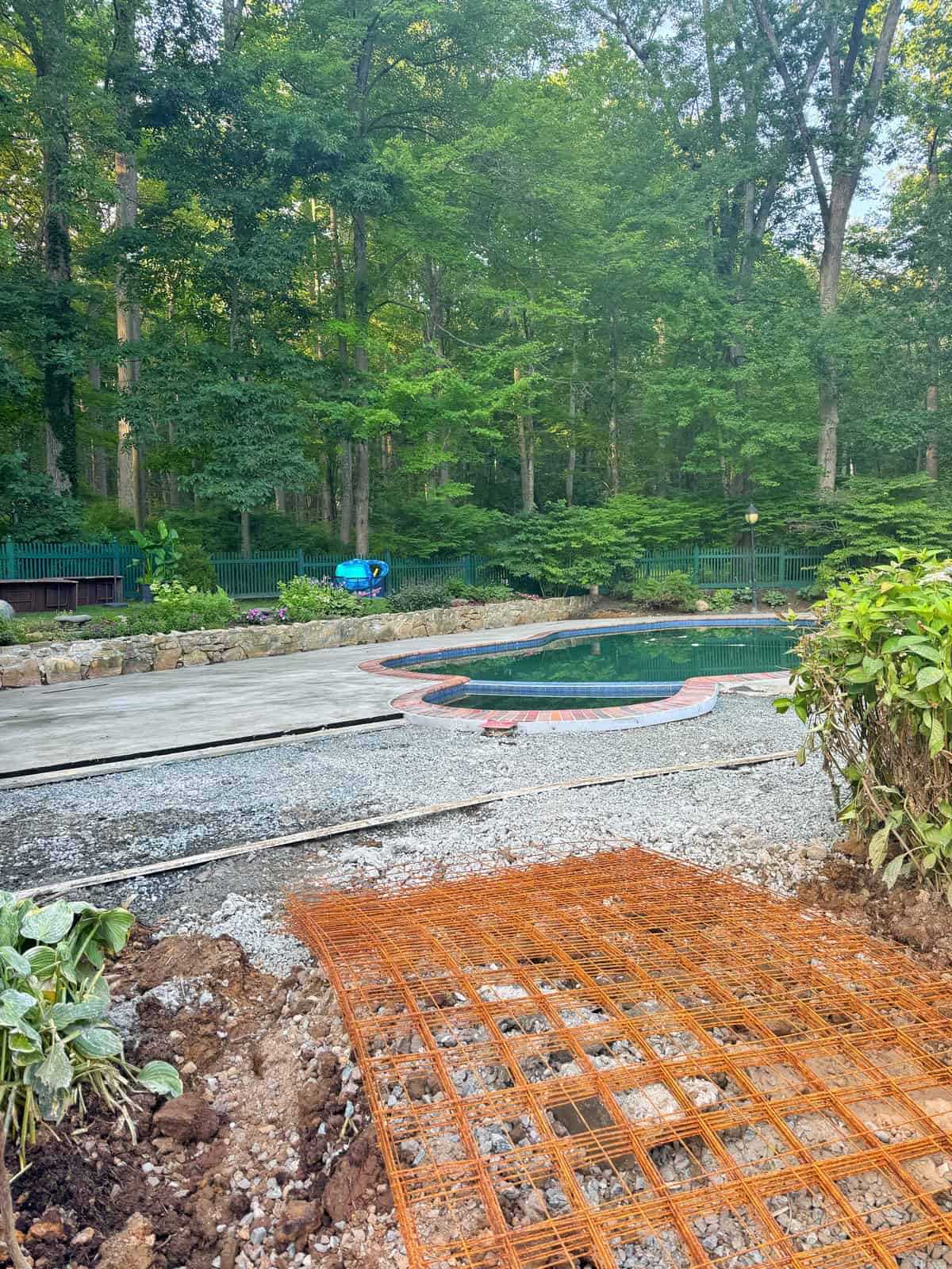 A backyard swimming pool surrounded by trees, with construction in progress. Gravel, orange metal rebar, and unfinished concrete are visible near the pool area, which is bordered by plants and a stone wall.