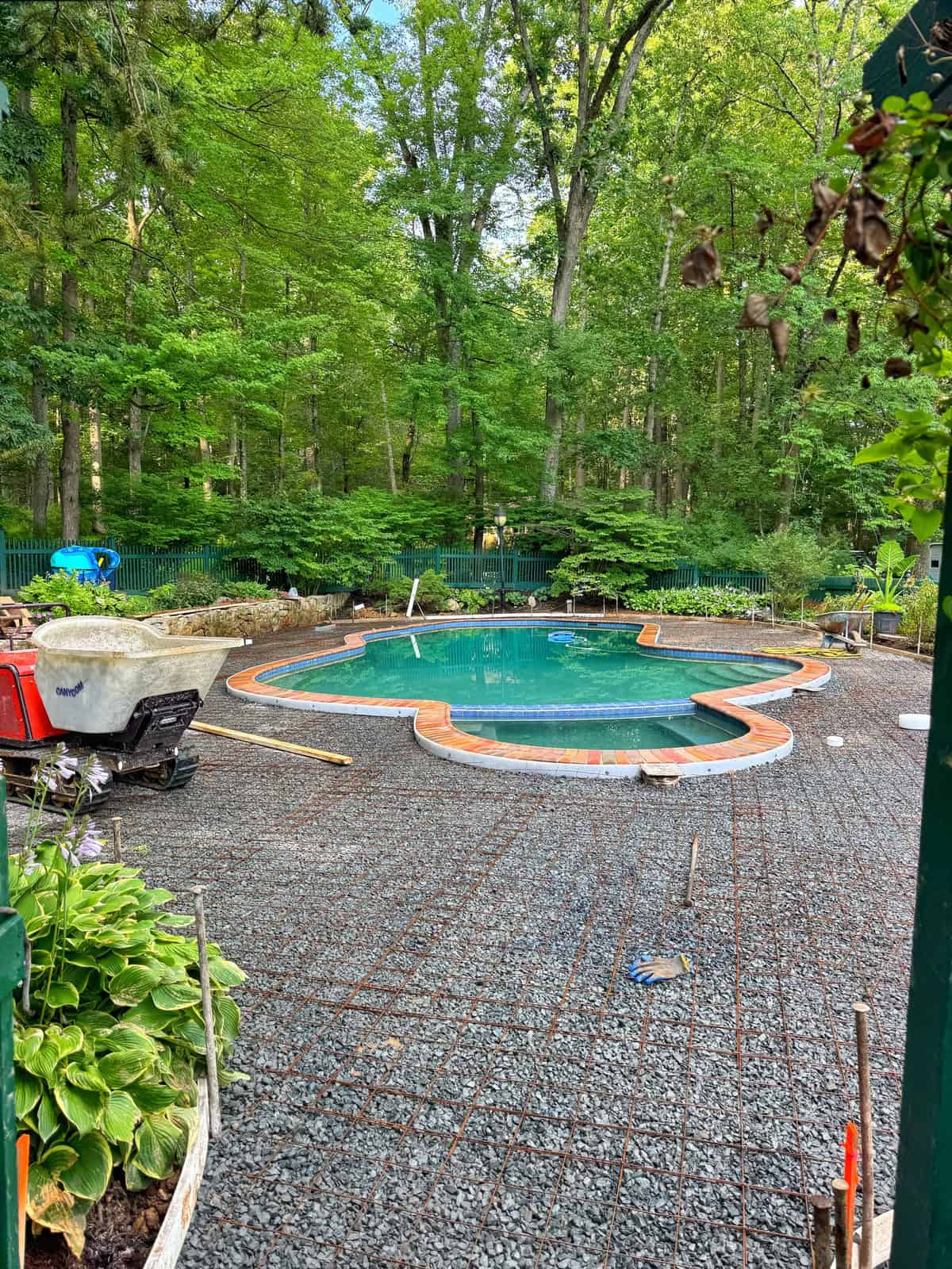 A partially finished backyard with a clover-shaped pool surrounded by gravel and construction tools, set in a lush, green wooded area. Rebar is laid on the ground, and landscaping work appears to be in progress.