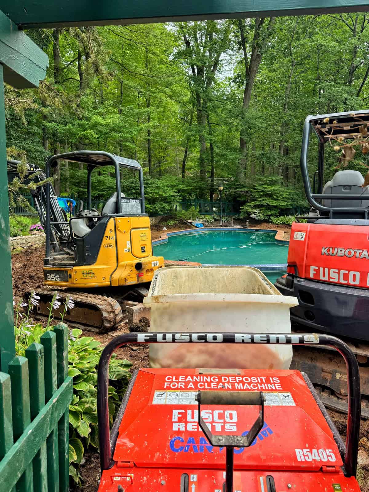 Excavators and construction equipment are parked near a backyard swimming pool, surrounded by trees and greenery, indicating ongoing landscaping or renovation work. The equipment displays "Fusco Rentals" branding.
