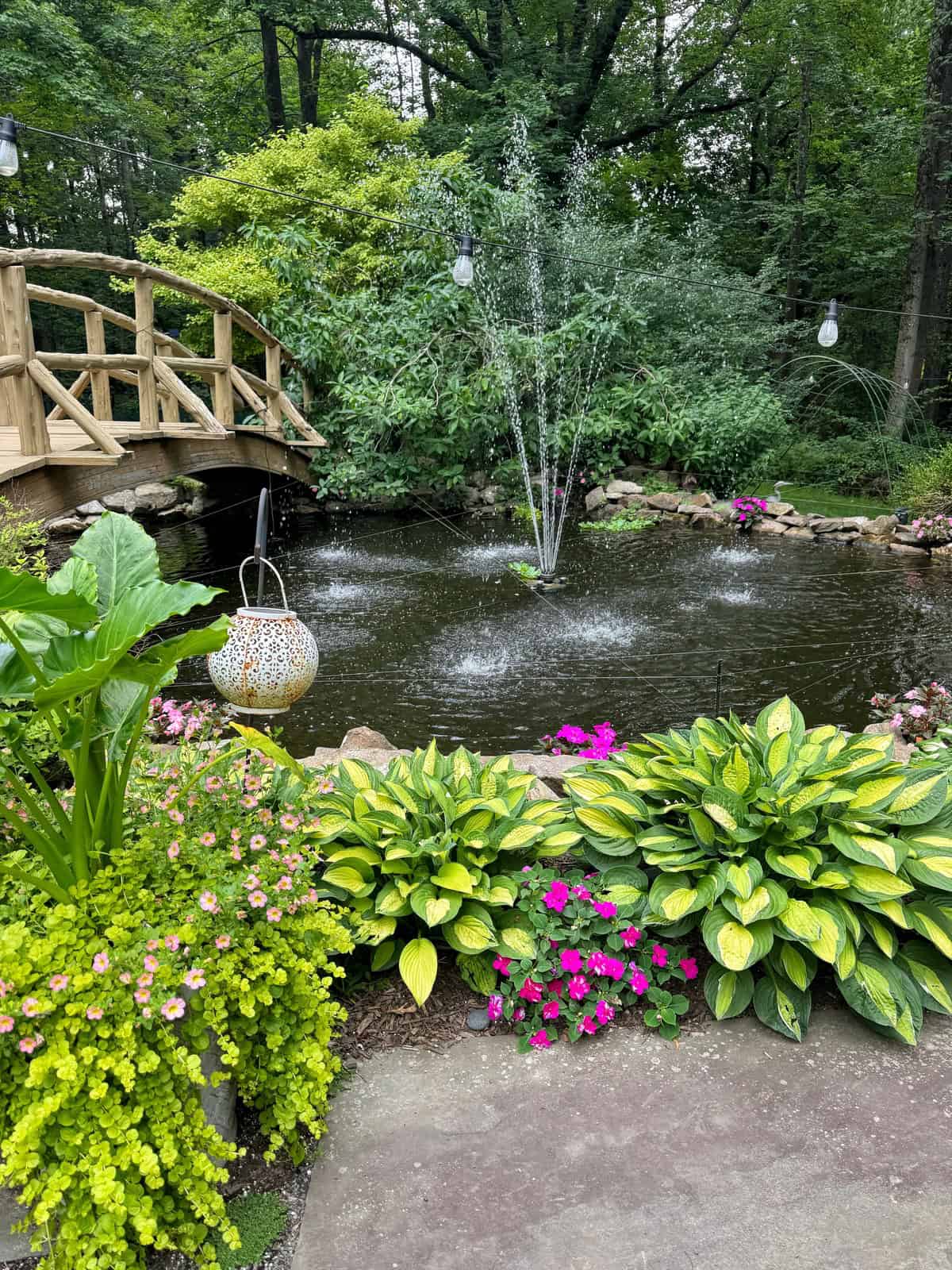 A garden pond with a small fountain, surrounded by lush green plants and pink flowers. A wooden bridge crosses the pond, and string lights hang above, creating a serene, inviting outdoor space.