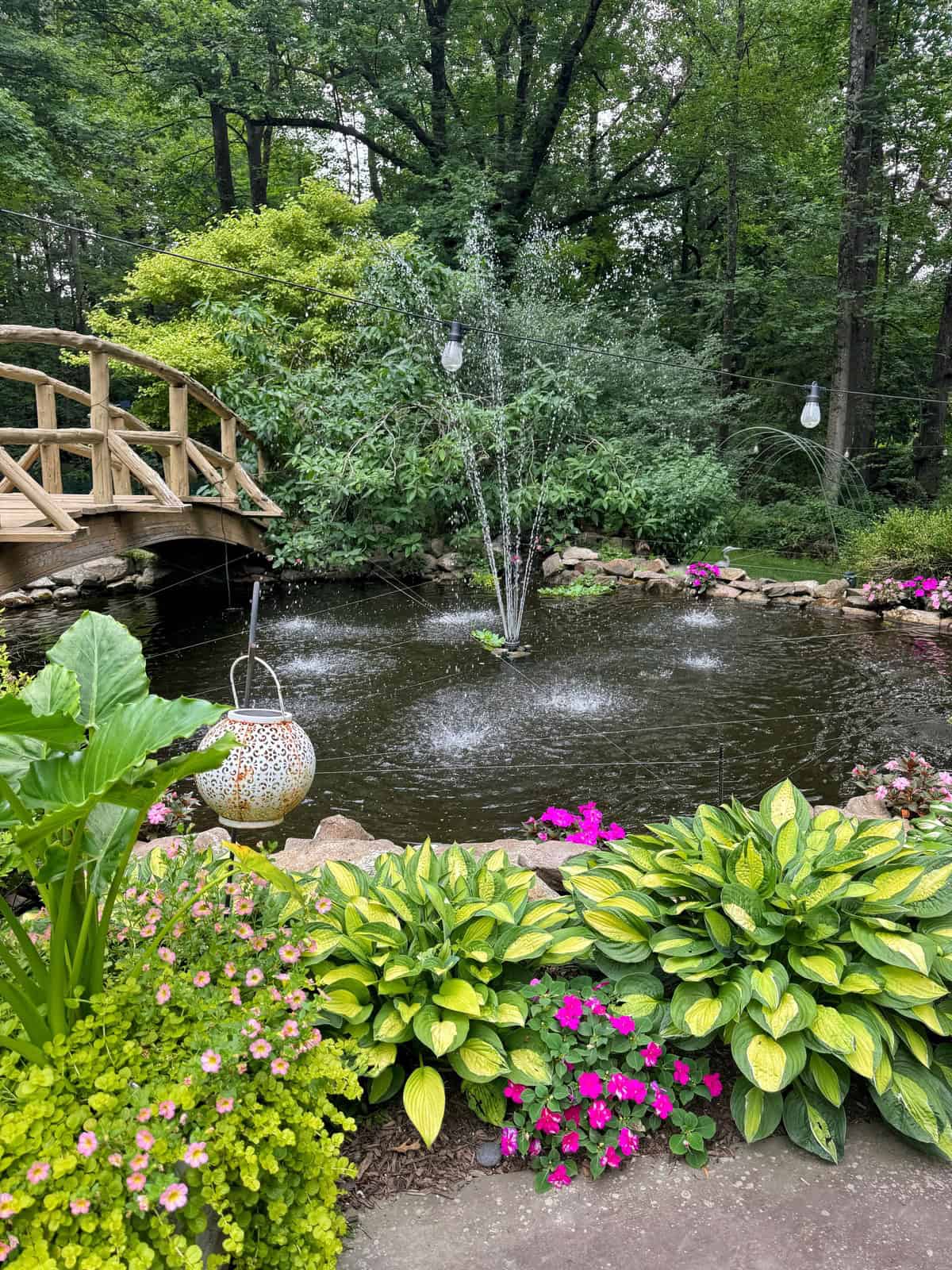 A lush garden pond with a fountain, surrounded by vibrant green plants and pink flowers. A wooden bridge spans one side of the pond, with hanging lanterns adding a decorative touch amidst the background trees.