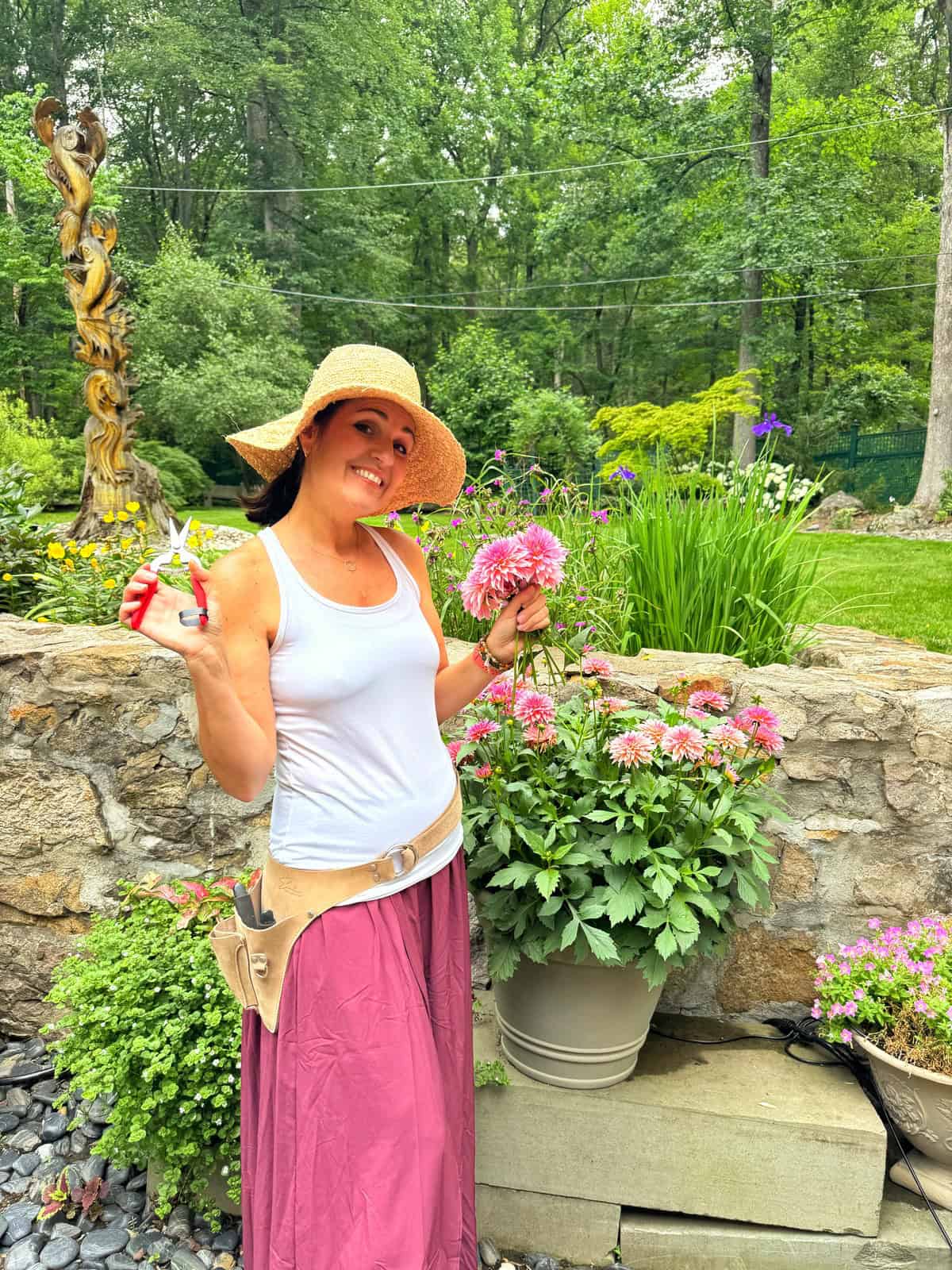 Stacy Ling is wearing a straw hat and long skirt stands in her zone 6b garden, smiling while holding pruning shears and pink dahlia flowers. She’s surrounded by blooming plants and greenery with a stone wall and sculpture in the background.