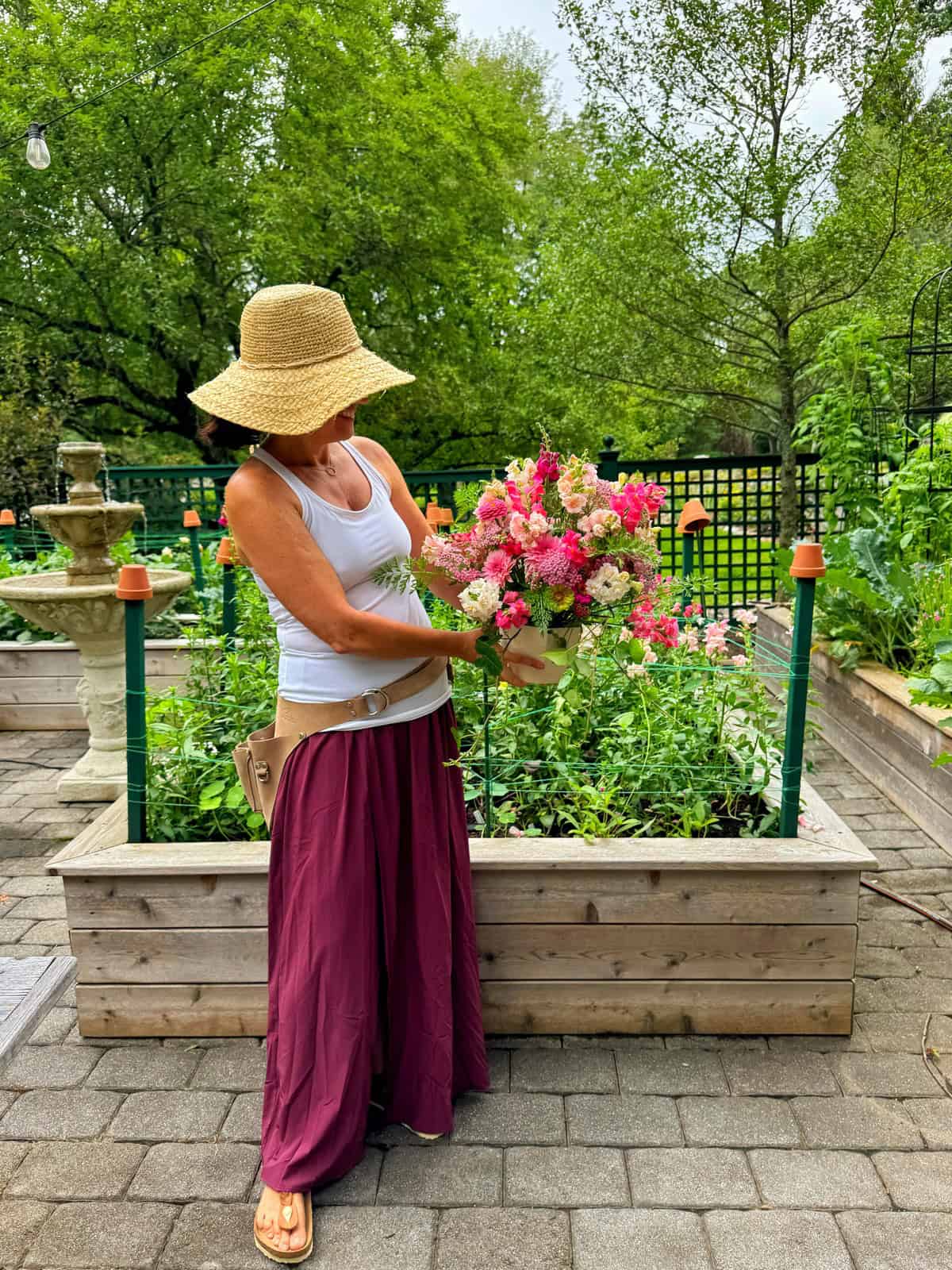 A woman (Stacy LIng) in a straw hat, white tank top, and maroon skirt stands on a garden path, holding a colorful bouquet of flowers, with raised garden beds and lush greenery in the background. Garden is located in zone 6b, New Jersey