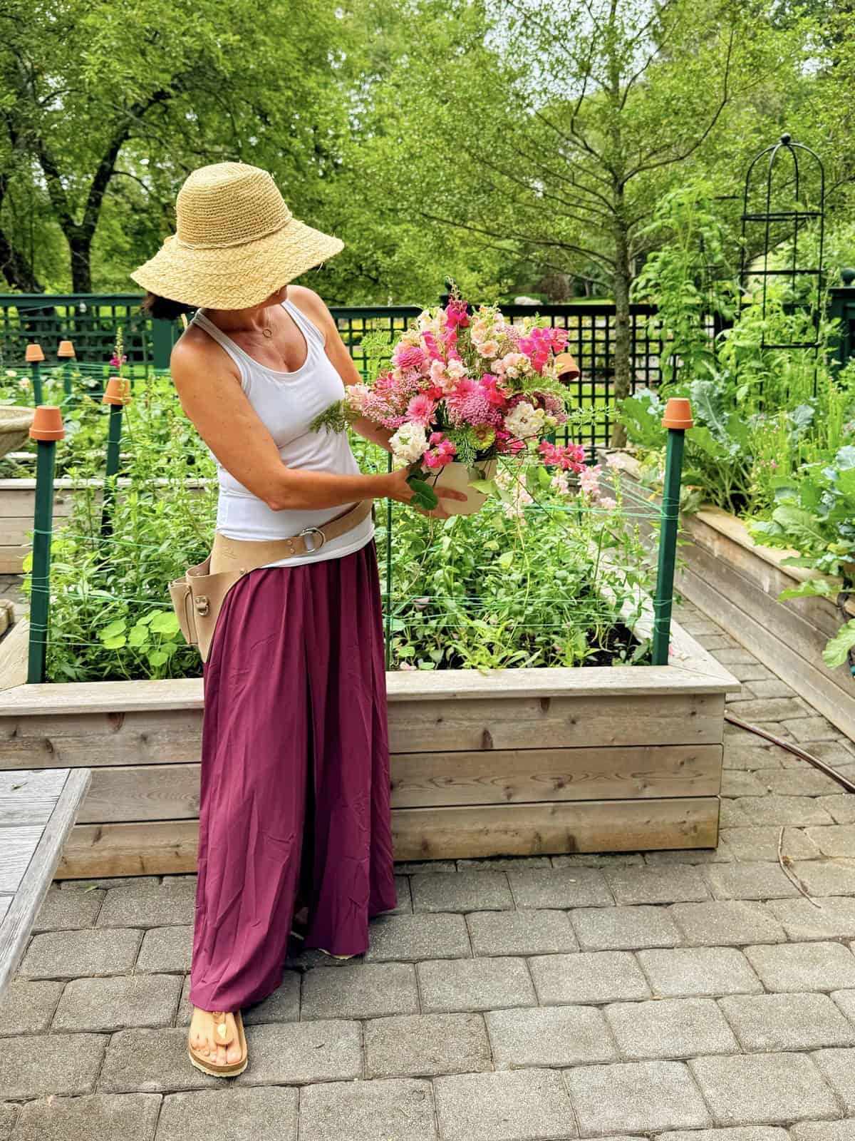 A woman in a straw hat, white tank top, and purple skirt stands on a garden path holding a bouquet of pink and white flowers. Raised garden beds with green plants and trees are in the background.