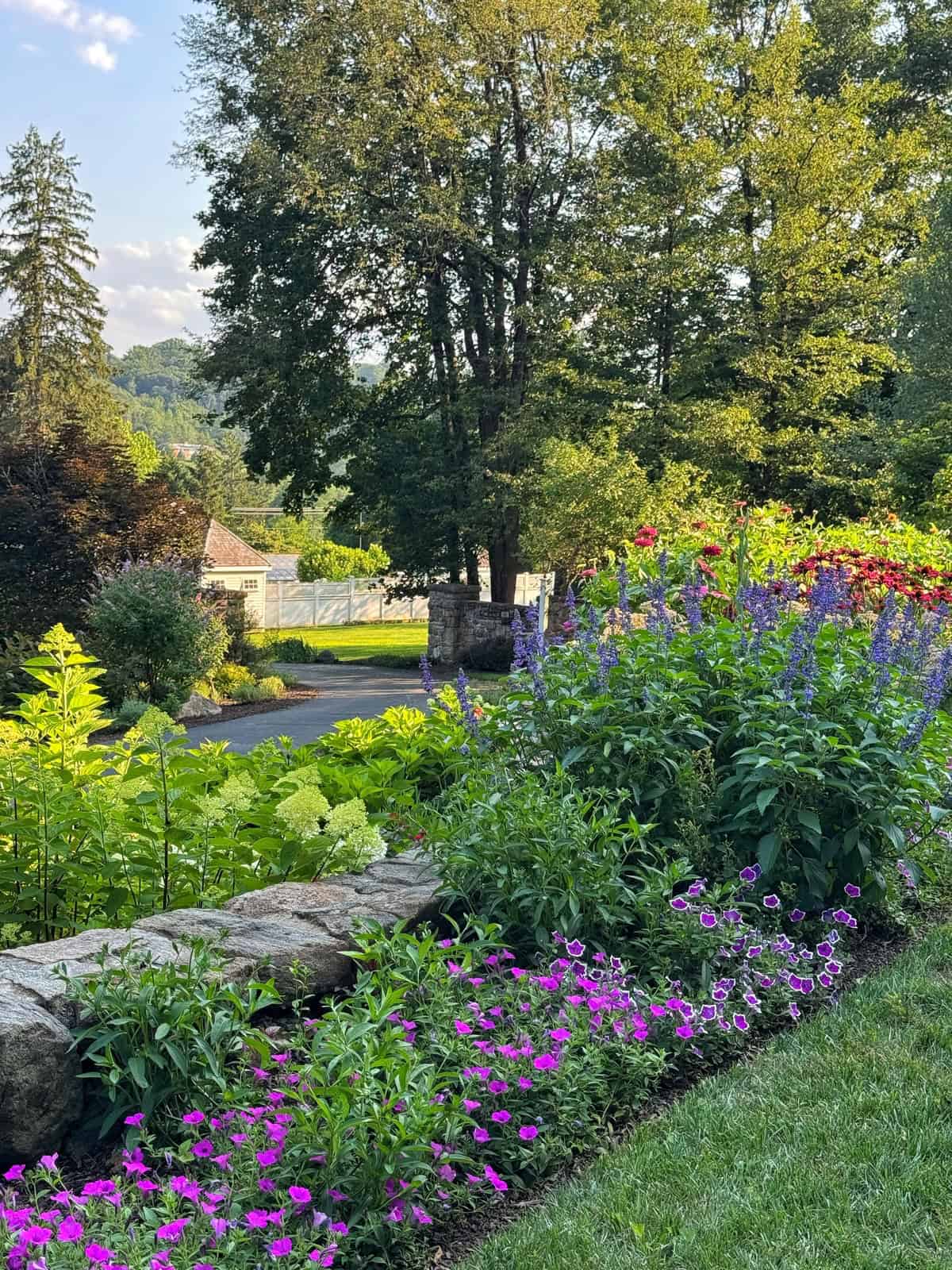 A lush garden with blooming pink, purple, and red flowers is bordered by a stone wall. Beyond the garden, there&rsquo;s a driveway, green trees, and a cottage-style building in the background under a blue sky.