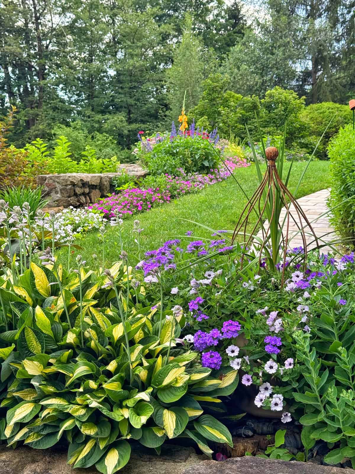 A lush garden with a variety of colorful flowers, green shrubs, and leafy plants, bordered by a stone path and surrounded by tall trees under a partly cloudy sky.