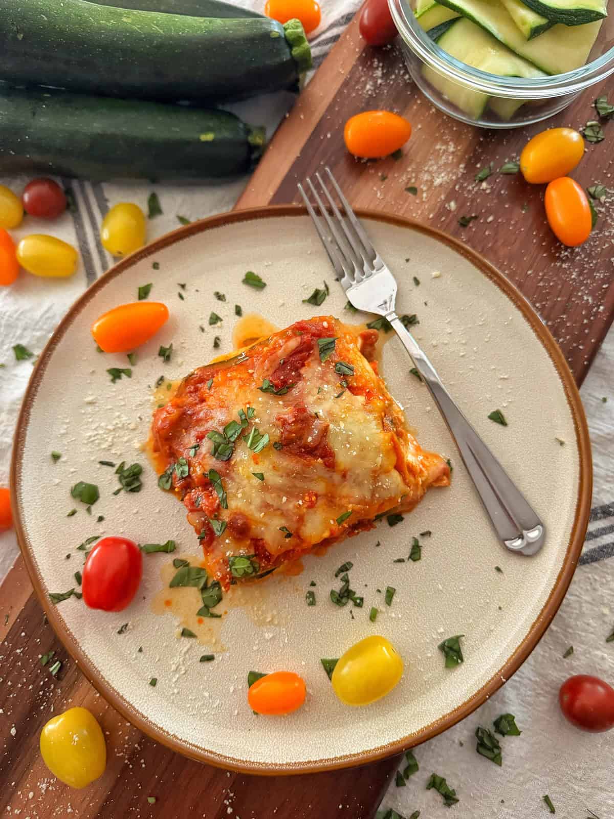 A plate with a serving of baked zucchini lasagna topped with melted cheese and herbs, surrounded by red and yellow cherry tomatoes. Zucchini and a bowl of zucchini slices are nearby on a wooden board. A fork rests on the plate.